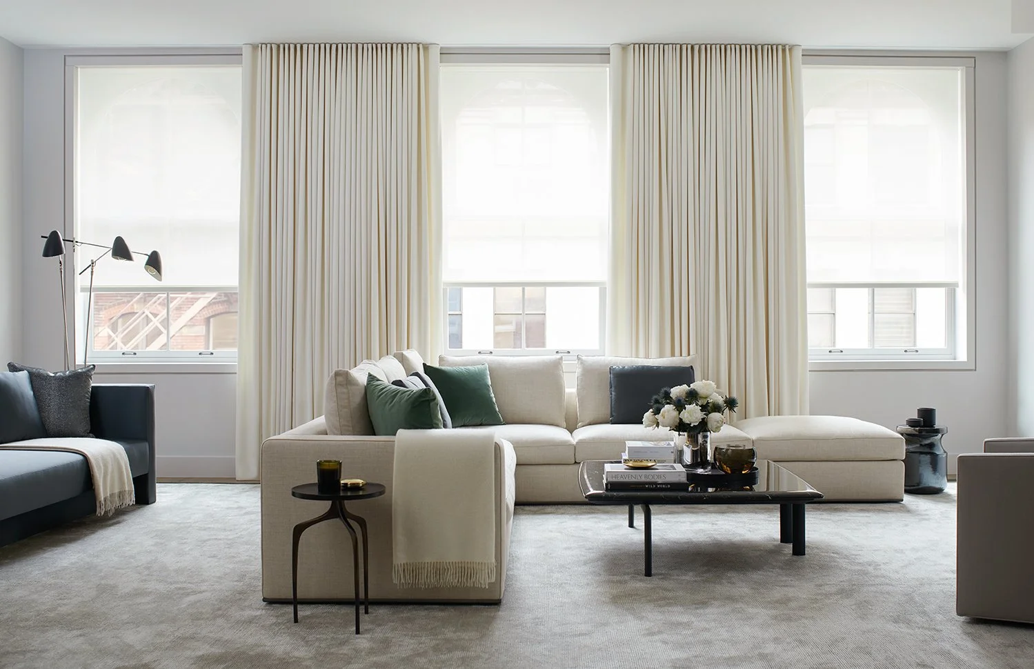 Bright living room with large windows, white curtains, beige sofa with green and navy pillows, modern black coffee table with books and flower arrangement, black side table with candle, black sofa with throw blanket, beige carpet, and minimal decor.