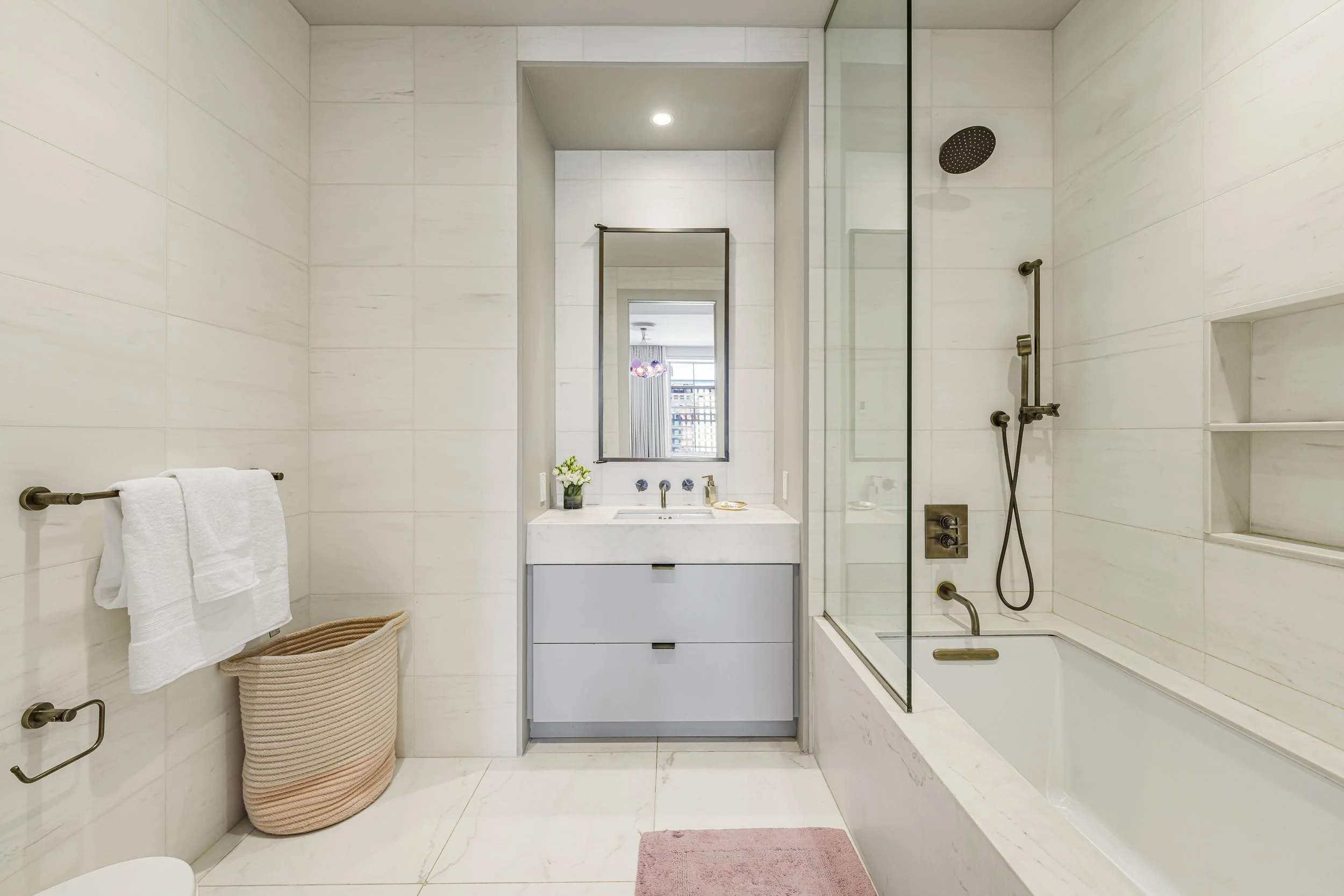 Modern bathroom with white tiled walls, a bathtub, a shower with a glass door, a vanity with a mirror, and a small pink rug on the floor.