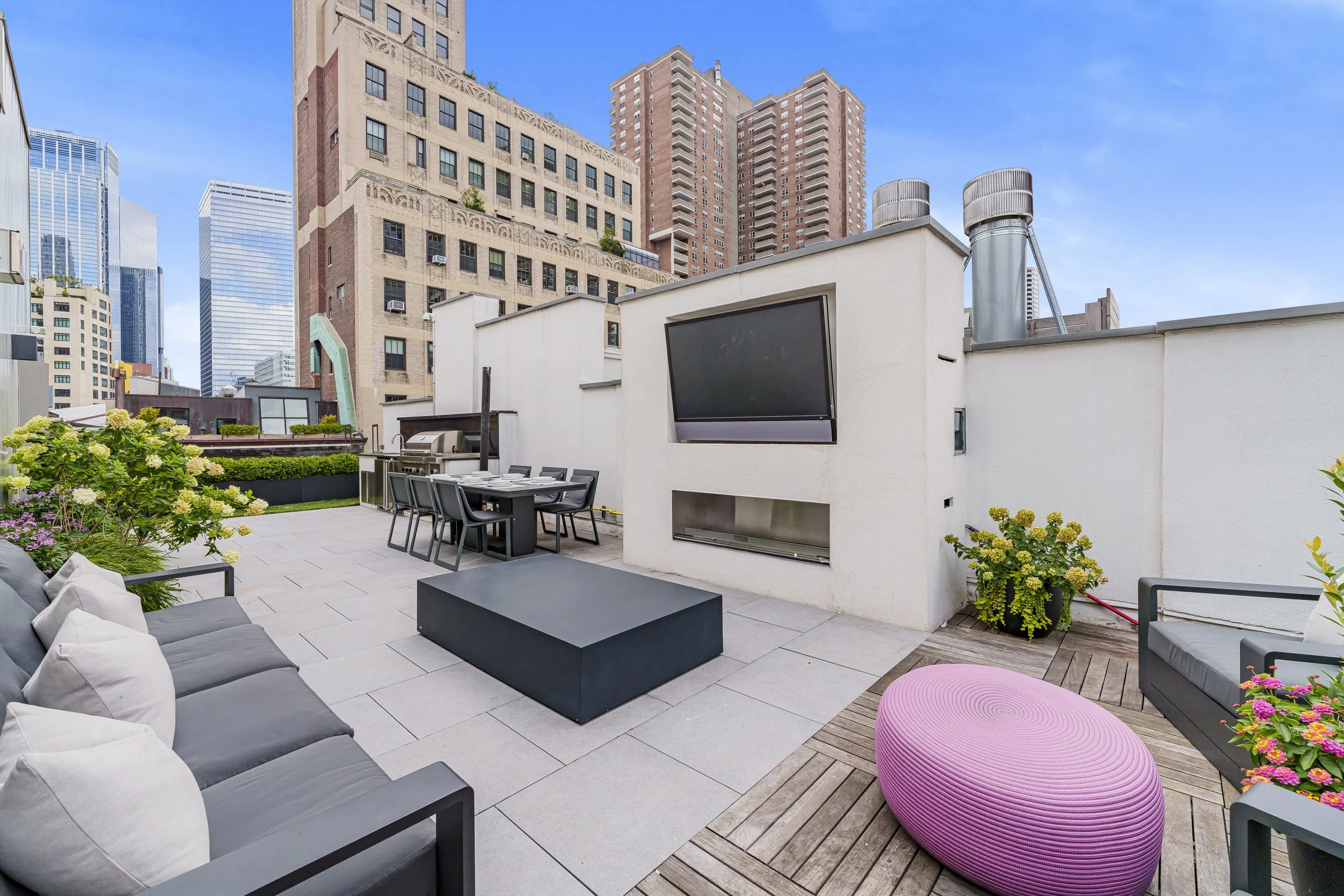 Rooftop patio with outdoor furniture, a large flat-screen TV, potted plants, and city skyscrapers in the background under a blue sky.