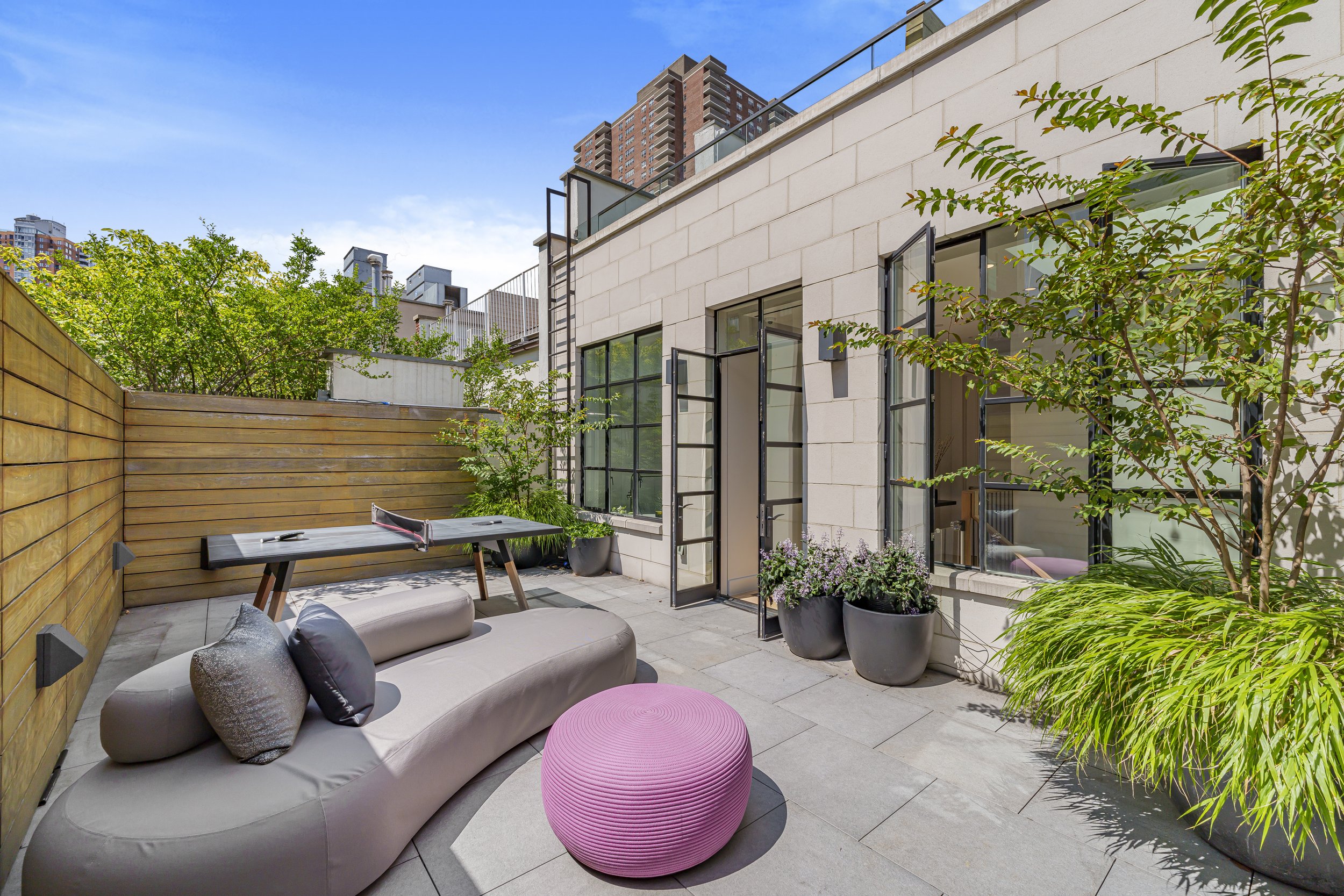 Modern outdoor patio with a beige curved sofa, pink ottoman, potted plants, and black foldable tables, surrounded by a wooden fence and adjacent to a white stone building with large windows and open black-framed doors, in an urban setting with tall b