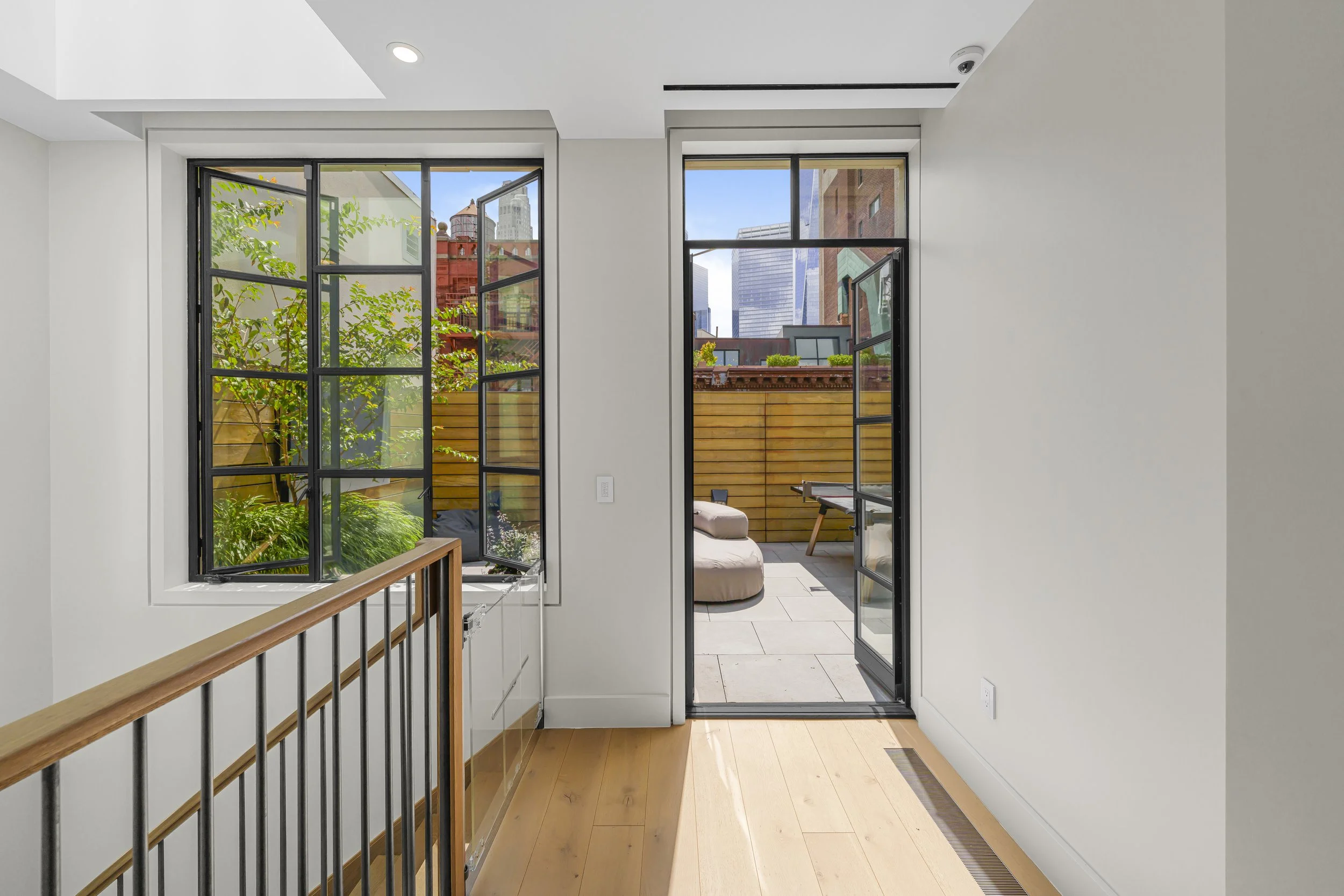 Interior view of a room with a wooden floor, white walls, a staircase railing on the left, an open door leading to a rooftop terrace, and a window showing outdoor greenery and city buildings.