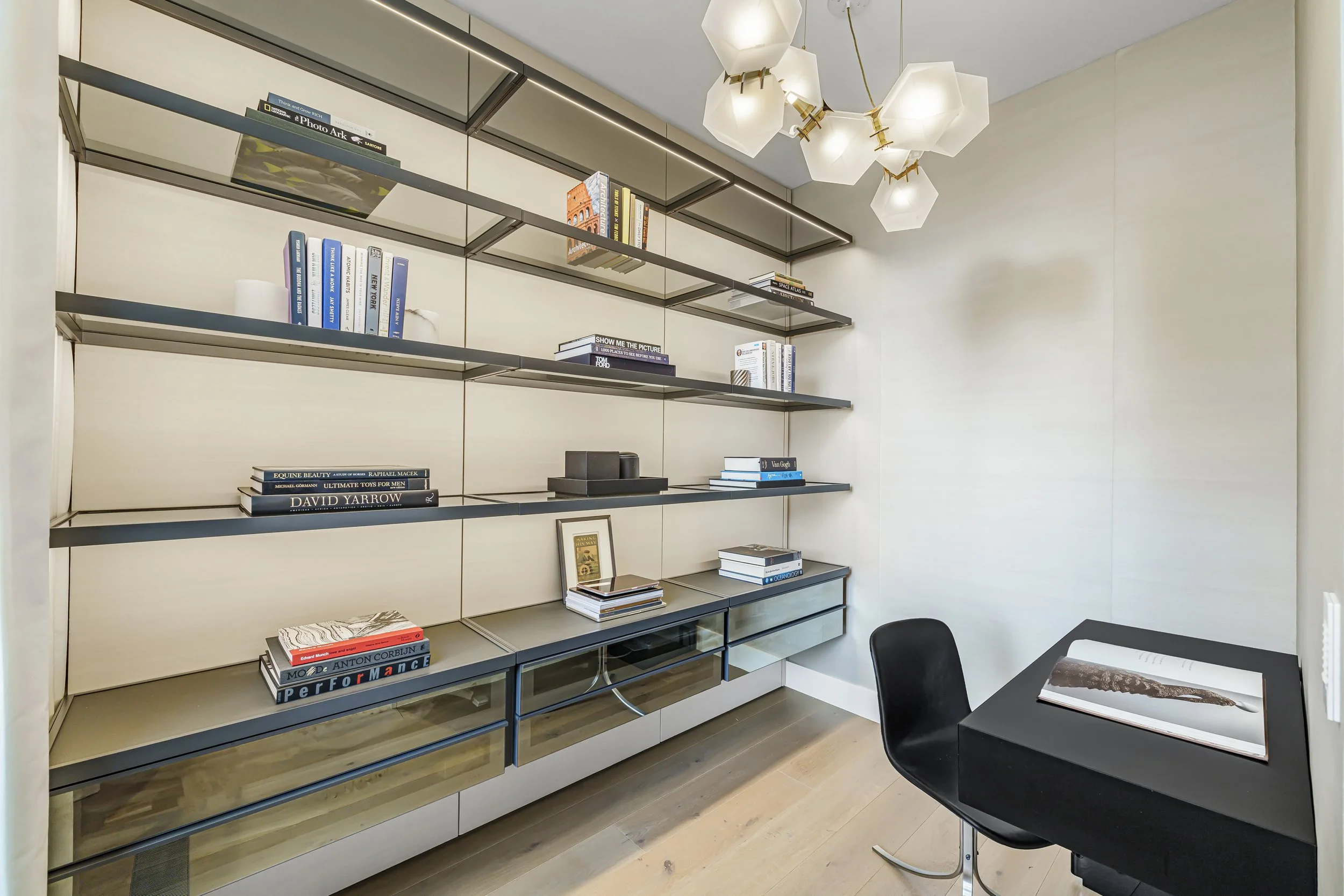 Modern home office with light-colored walls, a black desk with a chair, and a wall-mounted shelving unit filled with books and decorative objects.