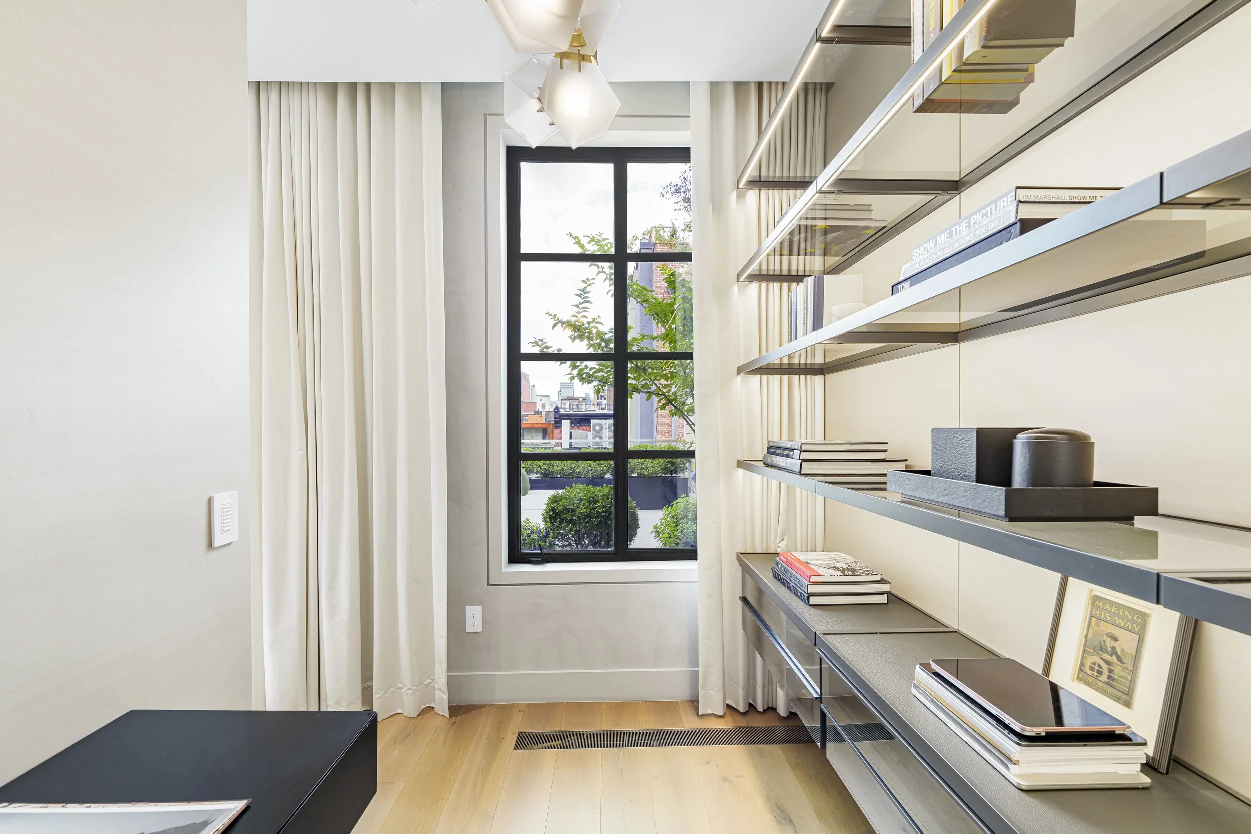 Interior view of a room with a large window, white curtains, a modern lighting fixture, and a shelving unit with books and decorative items.