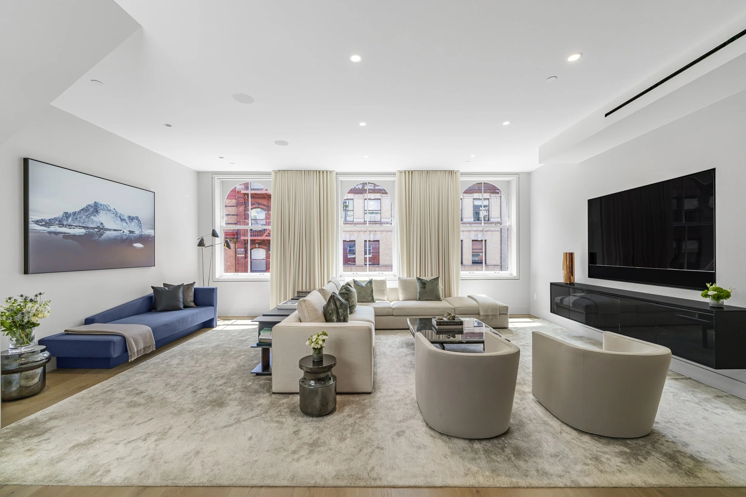 Bright, modern living room with white walls, large windows with cream curtains, a beige sectional sofa with throw pillows, two beige armchairs, a black TV mounted on the wall, and decorative plants and vases.