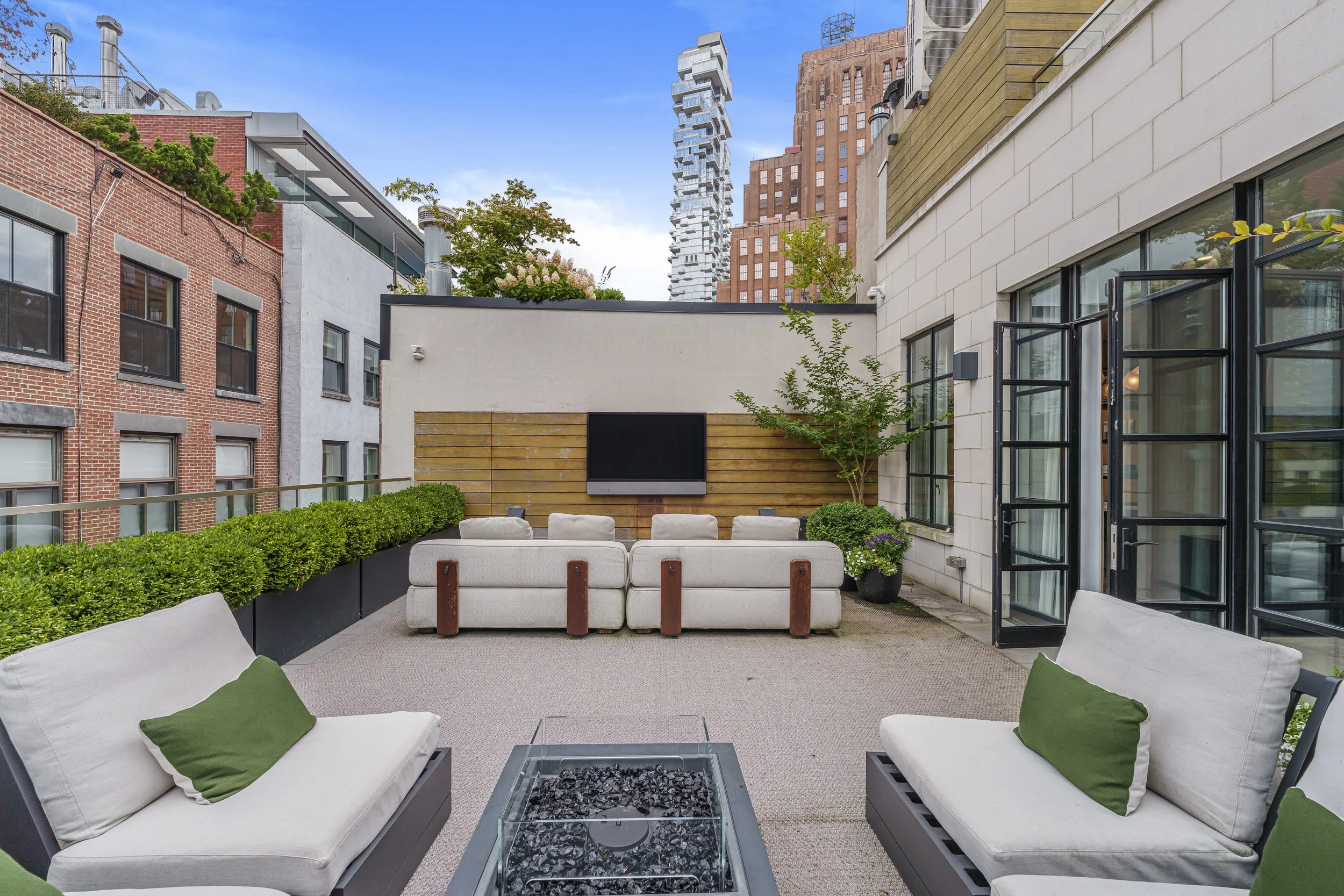 Outdoor rooftop patio with beige sofas, white cushions, green pillows, a fire pit, potted plants, an outdoor TV, and city buildings in the background.