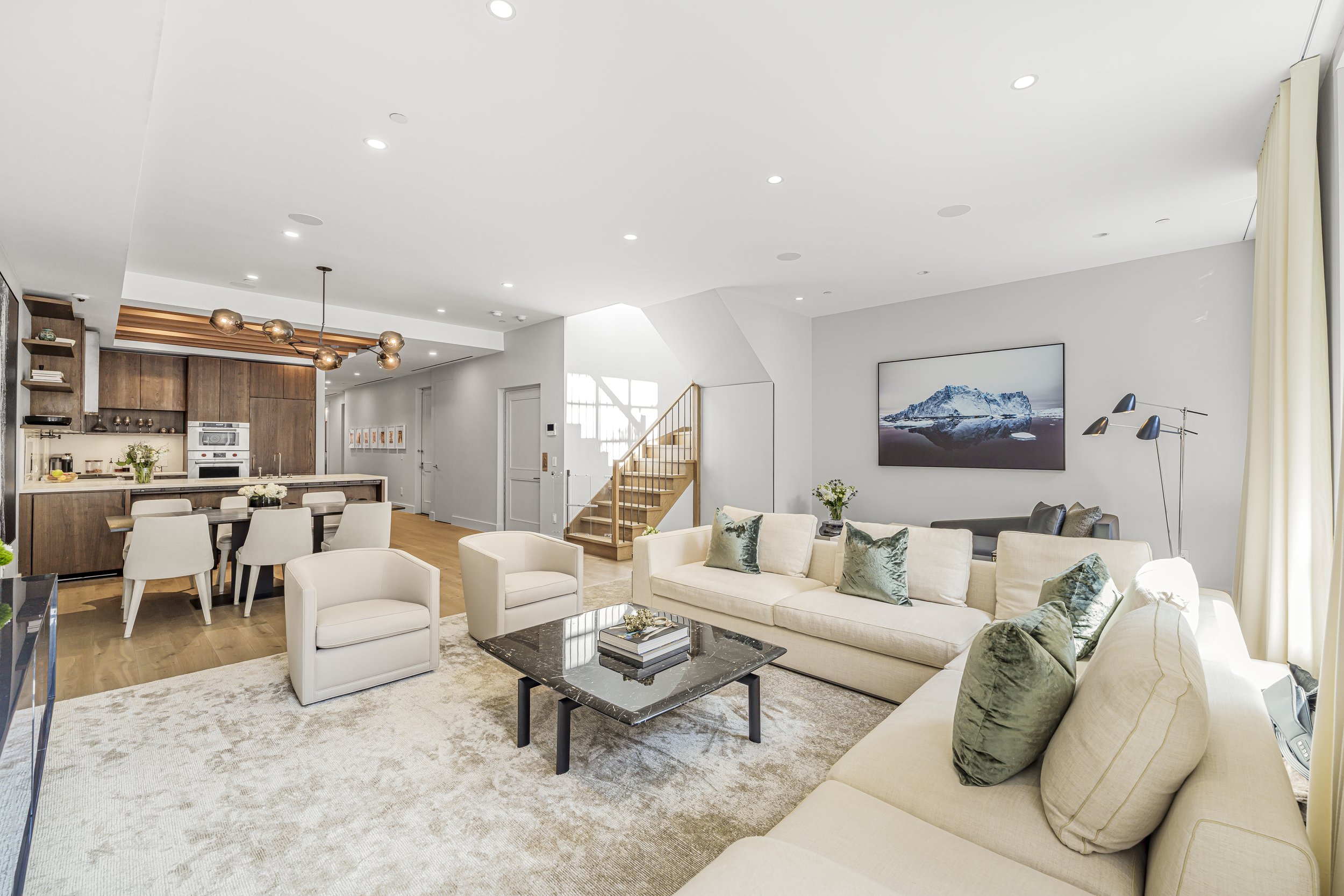 Living room with beige sofa and armchair, black marble coffee table, artwork of a mountain on wall, dining table with white chairs, kitchen in background with wooden cabinets, staircase with wooden steps, and large windows.