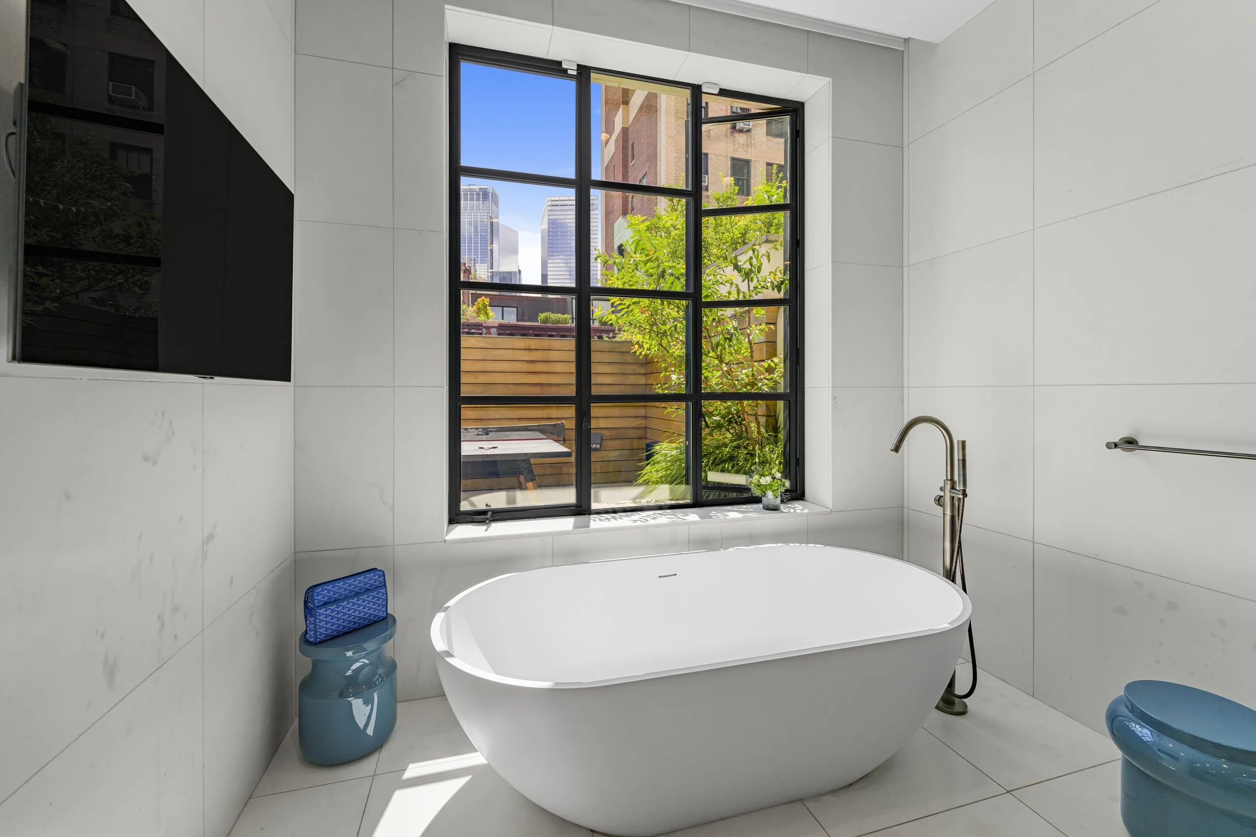 Modern bathroom with white tiled walls and floor, free-standing bathtub, large window with black frame showing city buildings and a small outdoor space with greenery, and blue bathroom accessories.