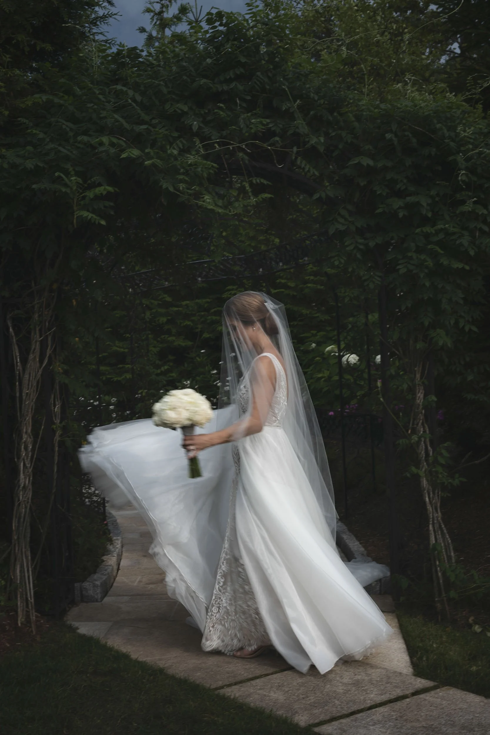 A bride takes a twirl in her wedding dress near a canopy of green trees.