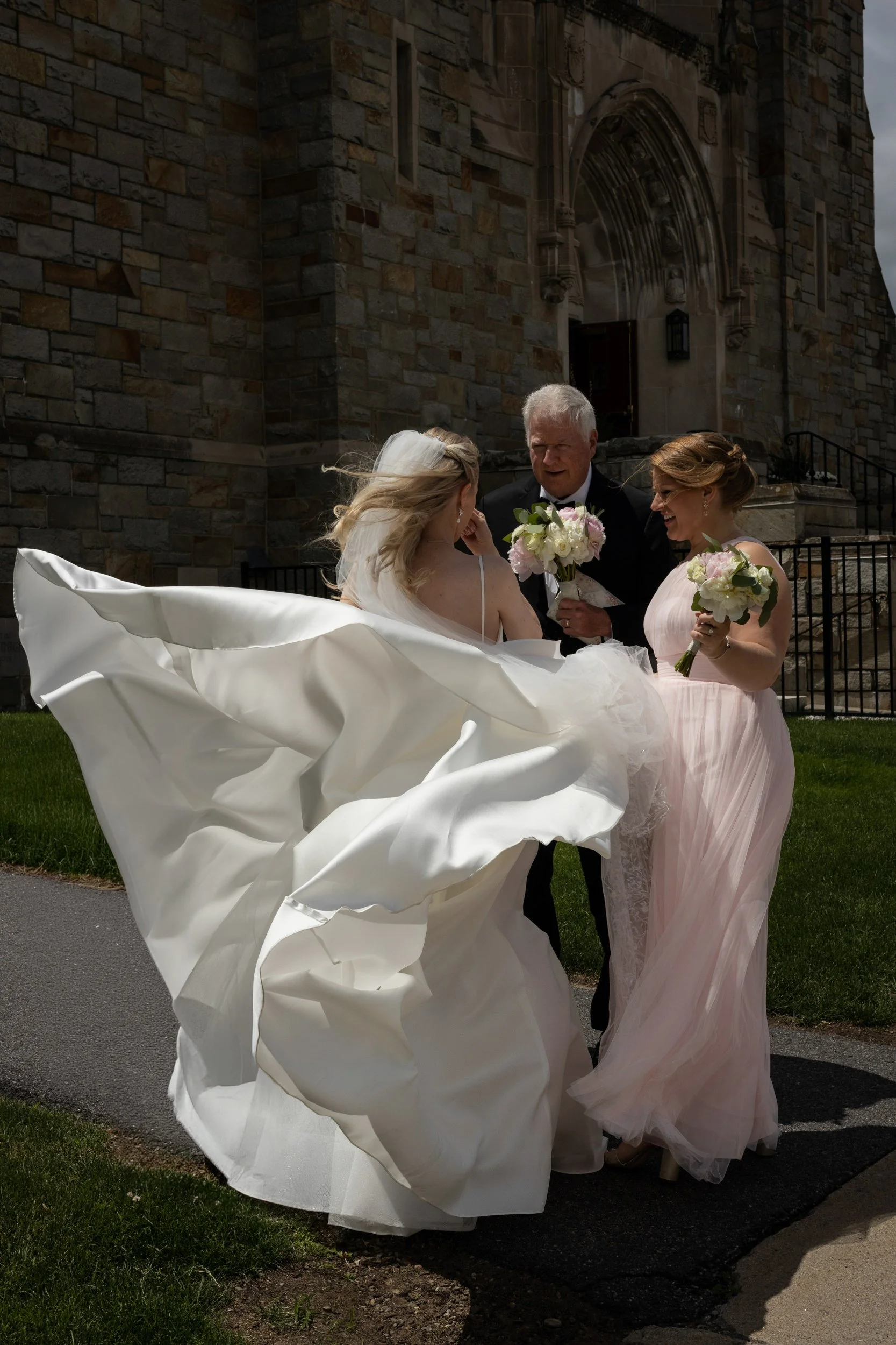 Windy wedding day as the bride stands outside the church with her father and sister before going inside.