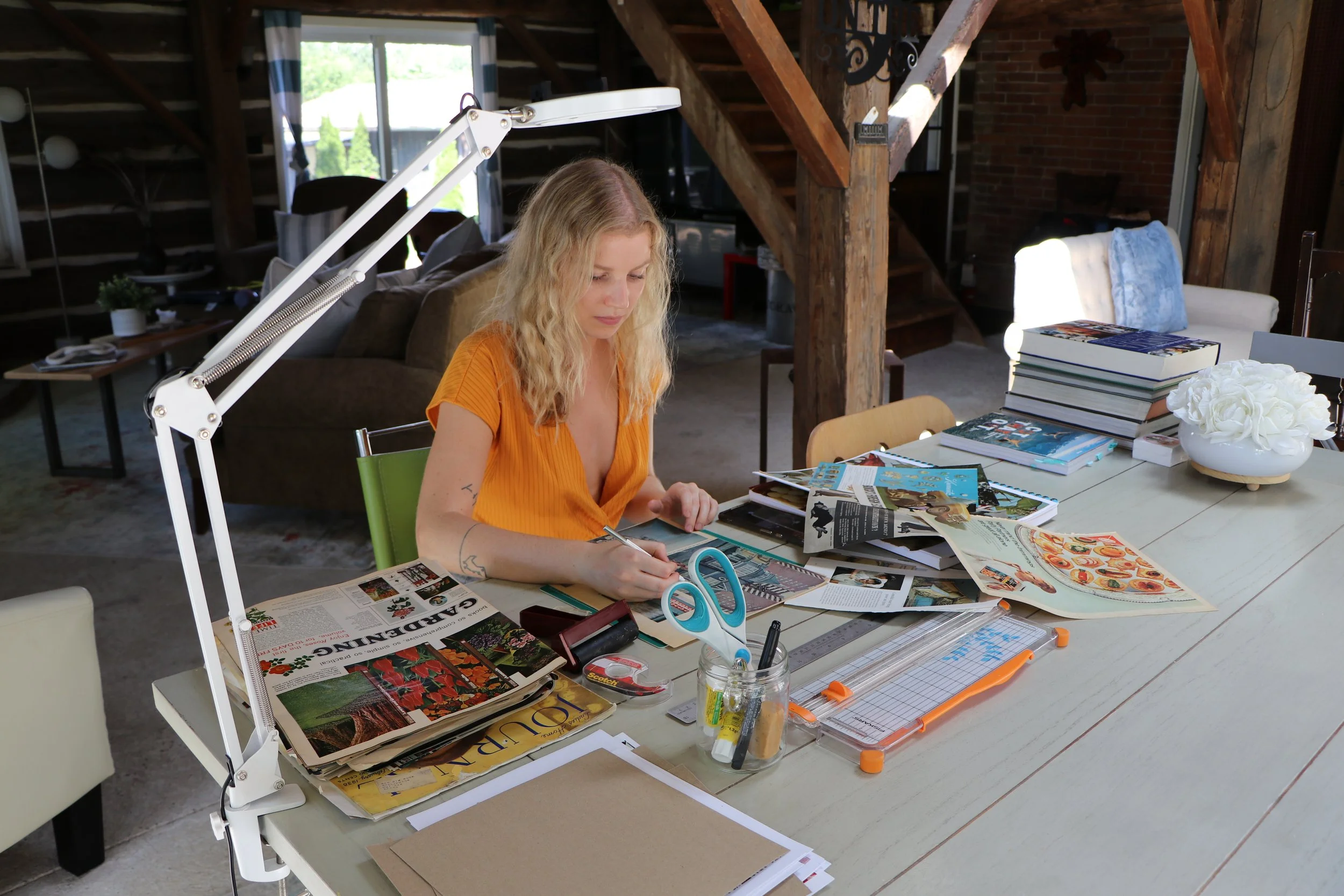 A woman with blonde hair wearing an orange shirt sitting at a cluttered table with magazines, papers, scissors, and art supplies, working on a creative project in a rustic room with wooden beams and a staircase.
