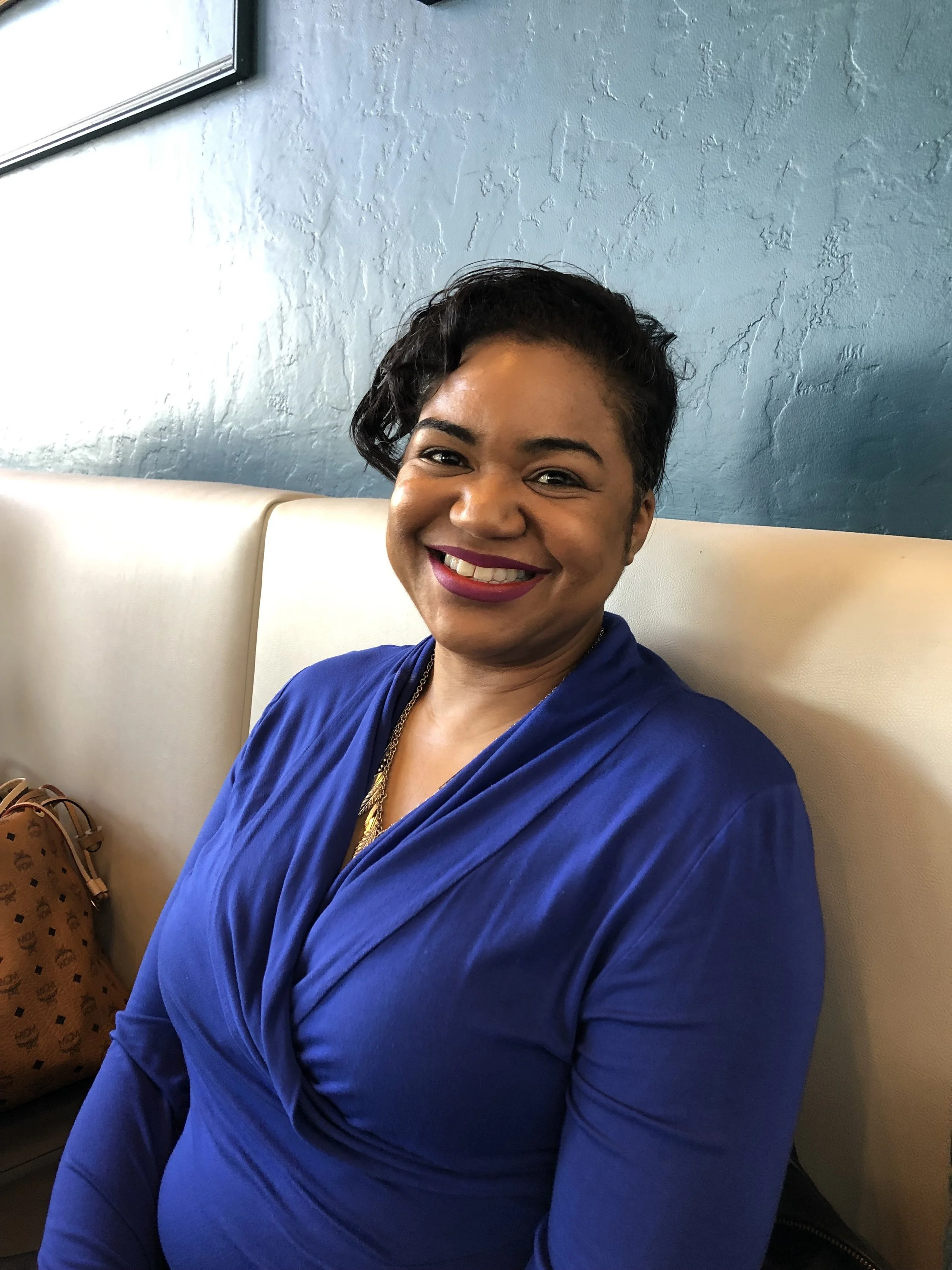 Smiling woman in blue dress seated indoors, with textured wall background.