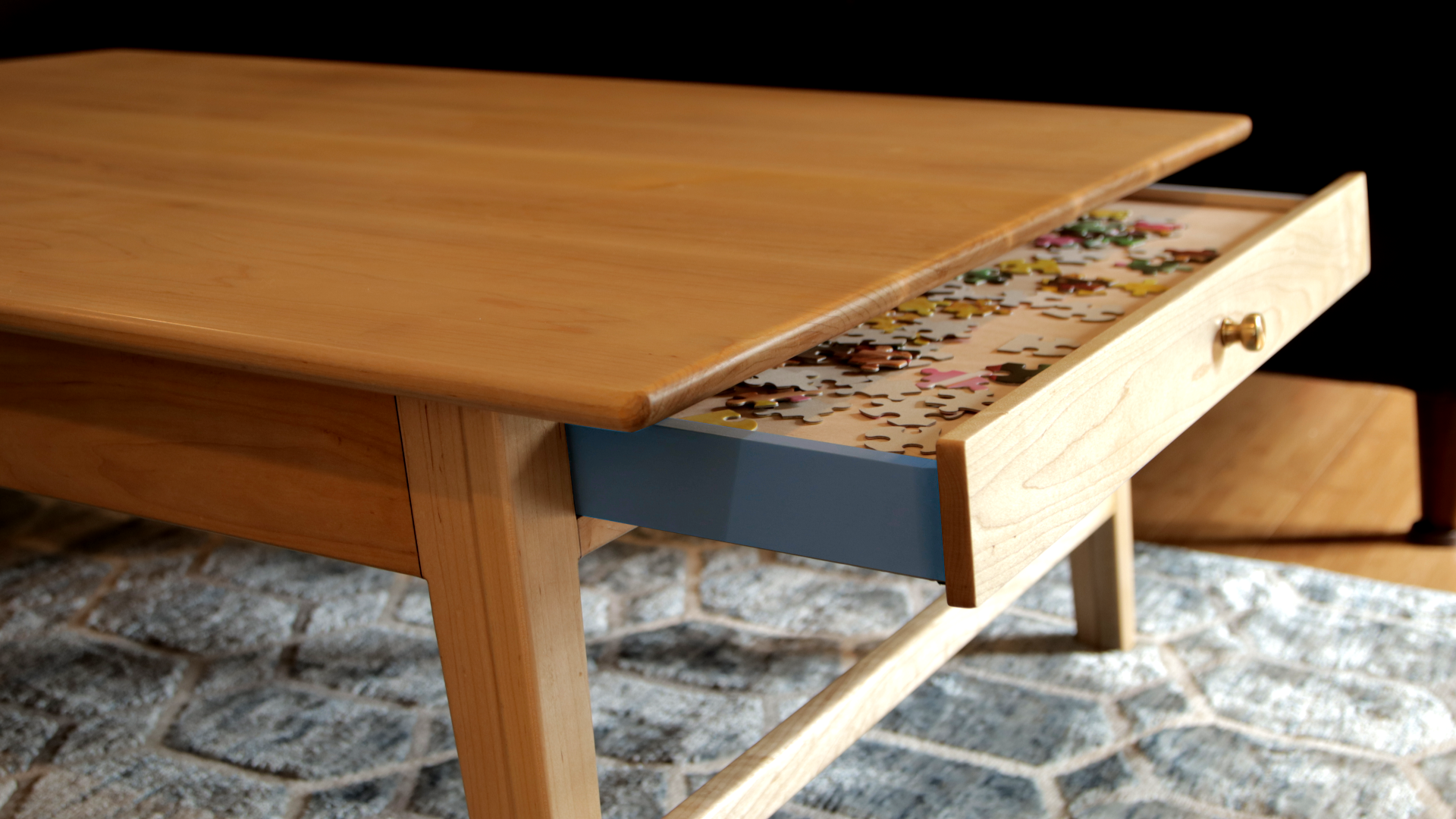 Three-quarter view of danish modern coffee table. The table is made of maple with walnut accents. There is a drawer that is partially pulled out. The sides of drawer are blue in color. Inside the drawer are puzzle pieces.