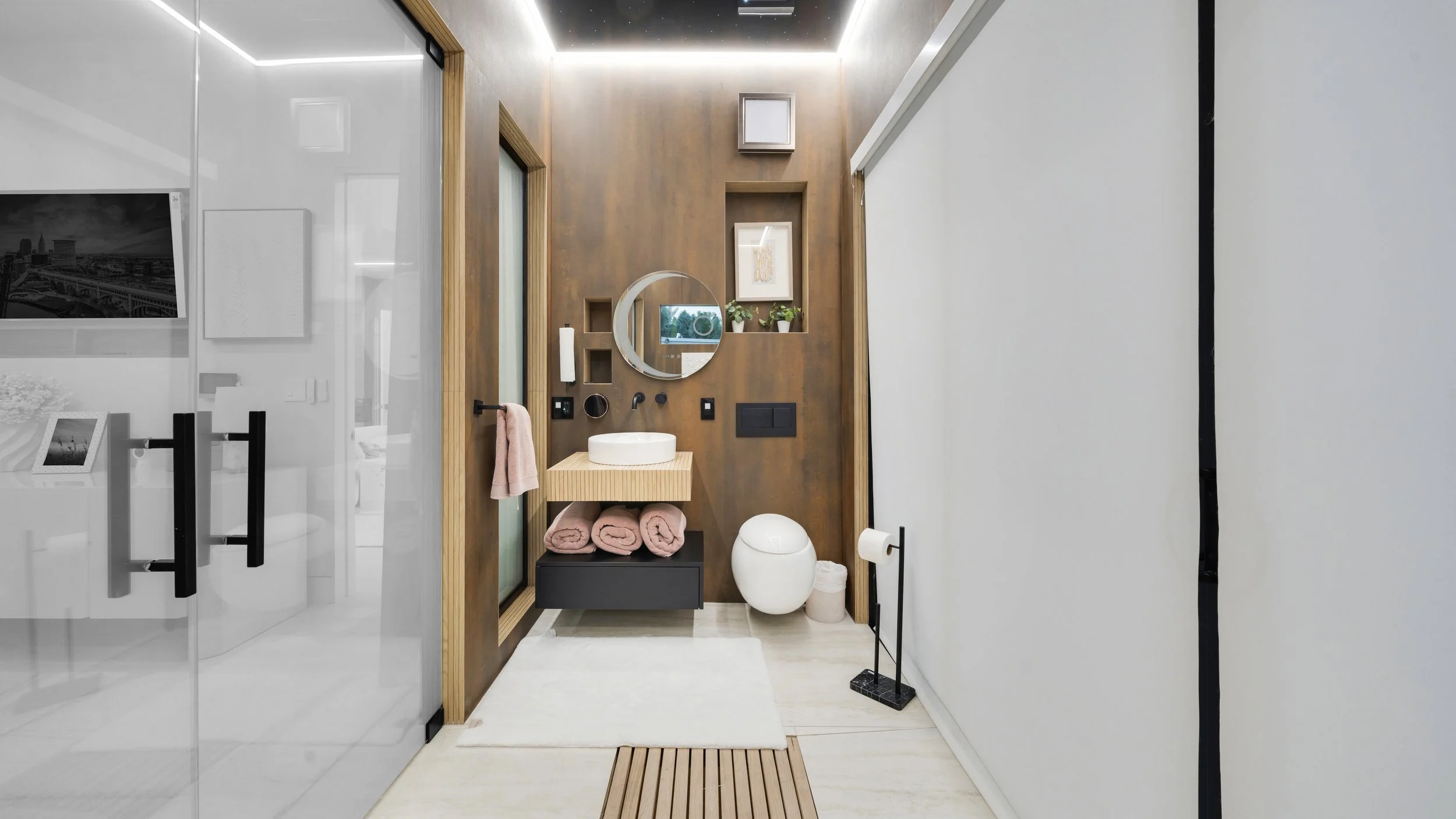 Modern bathroom with wood-paneled accent wall, round mirror, small vessel sink, black drawer with pink towels, white toilet, towel holder with pink towel, potted plants, framed artwork, and minimalist decor.