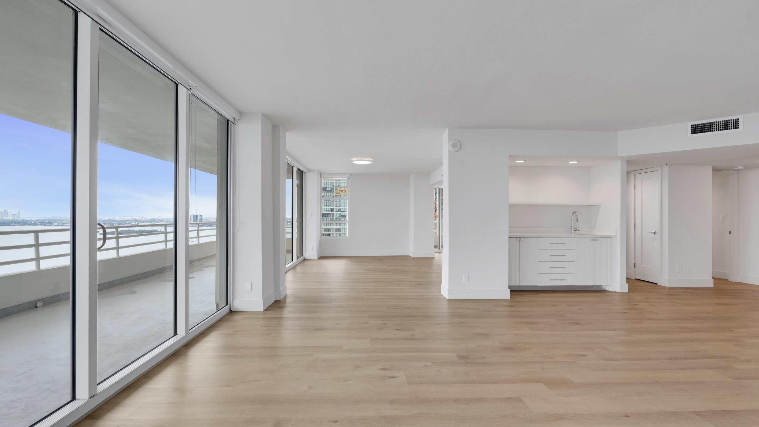 Empty modern apartment with large floor-to-ceiling windows and a balcony, white walls, light wood flooring, and a small white kitchen area.