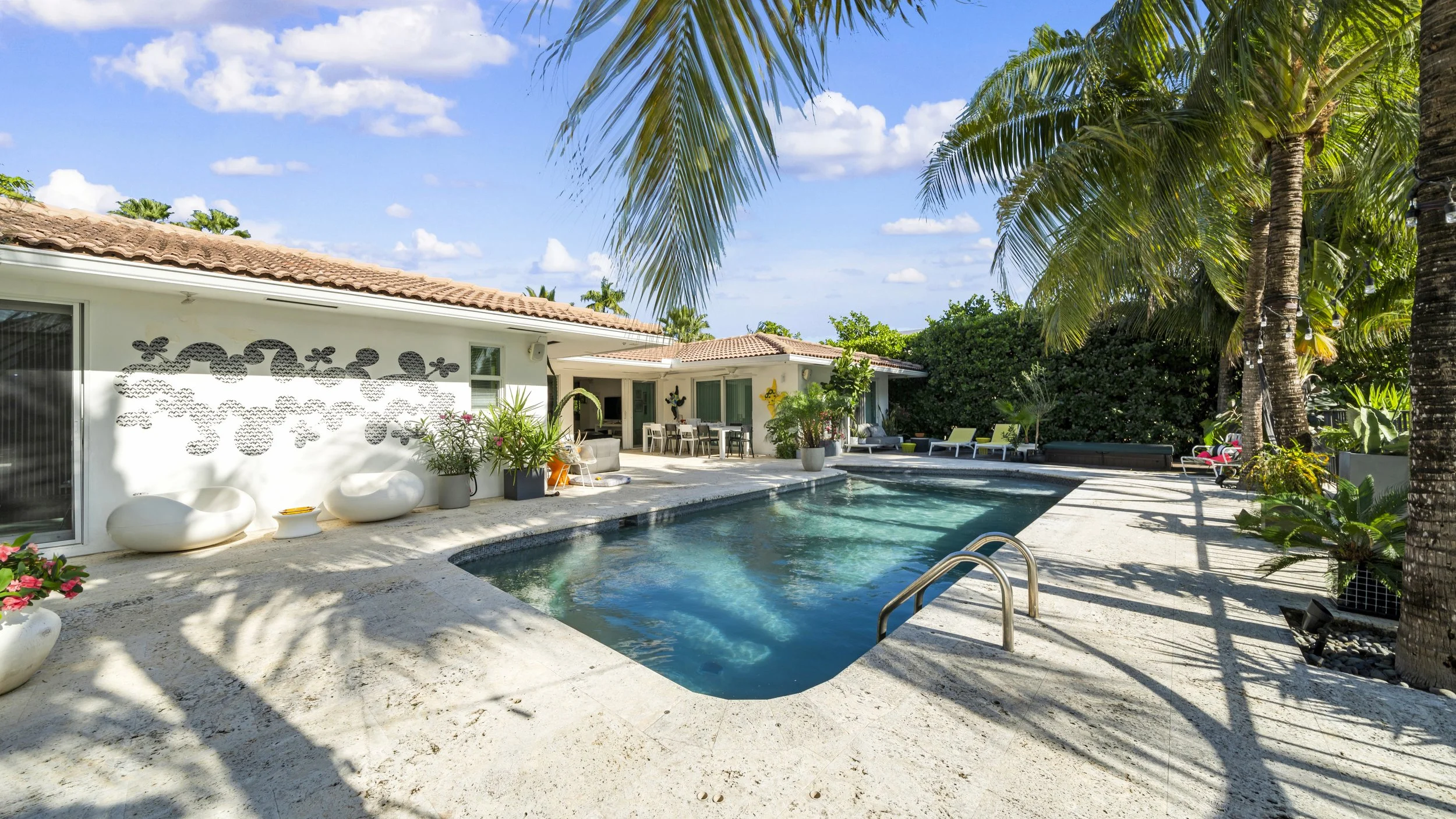 A backyard with a swimming pool, surrounded by palm trees and tropical plants, with part of a white house and outdoor furniture visible under a clear blue sky.