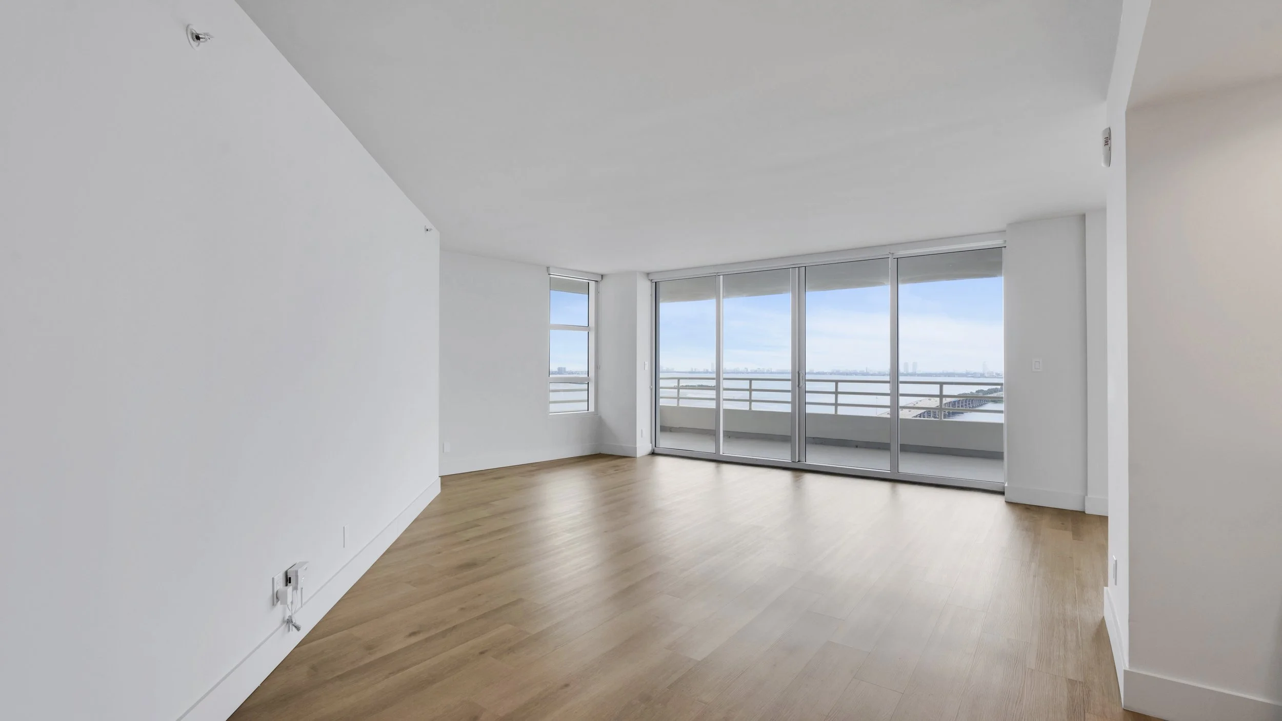 Empty living room with large windows and sliding glass doors leading to a balcony, with city skyline view in the distance, light wood flooring, white walls, and ceiling.
