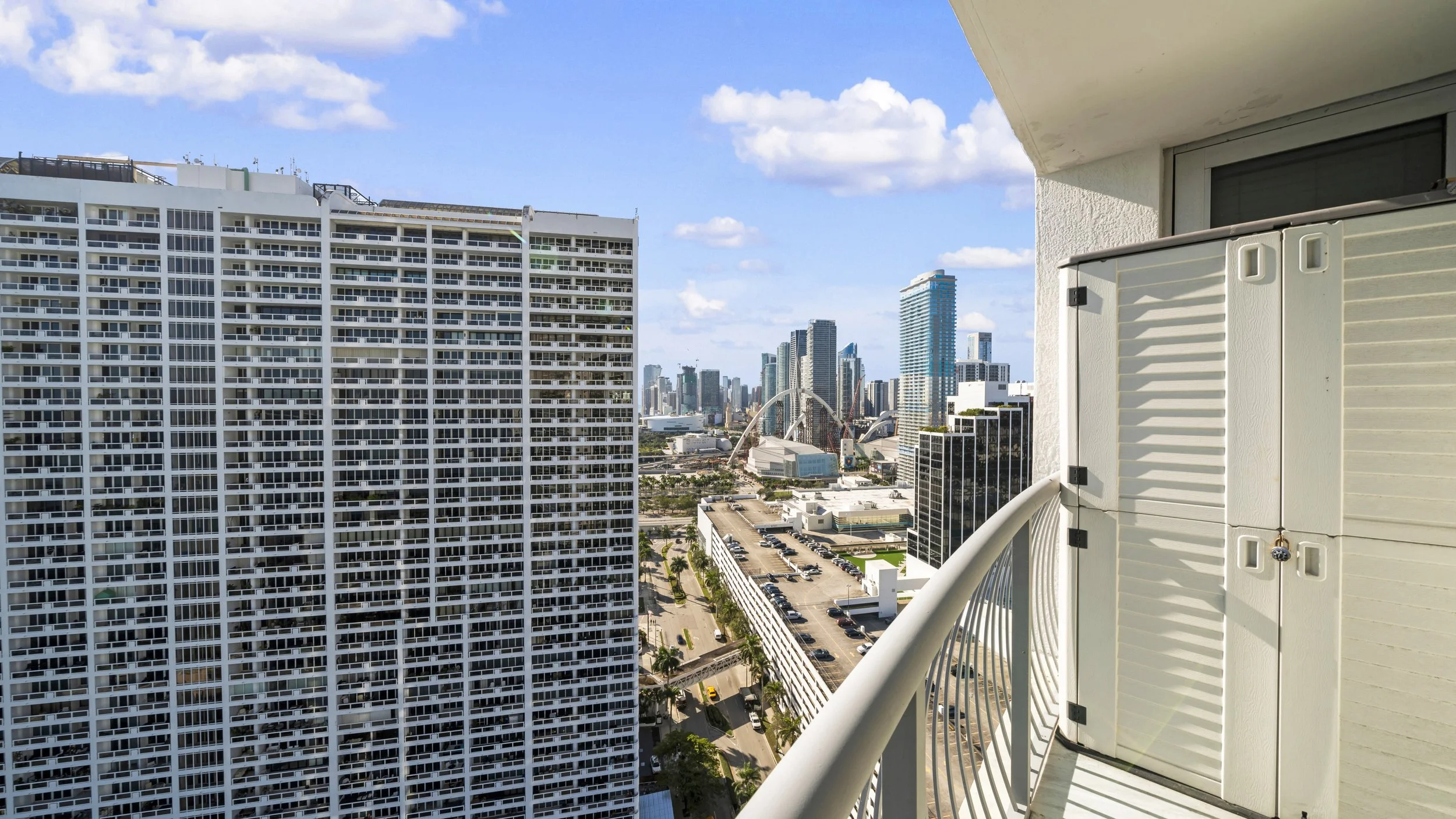 City view from a balcony with a high-rise building in the foreground, a parking lot below, and a skyline with tall skyscrapers and a clear blue sky in the background.