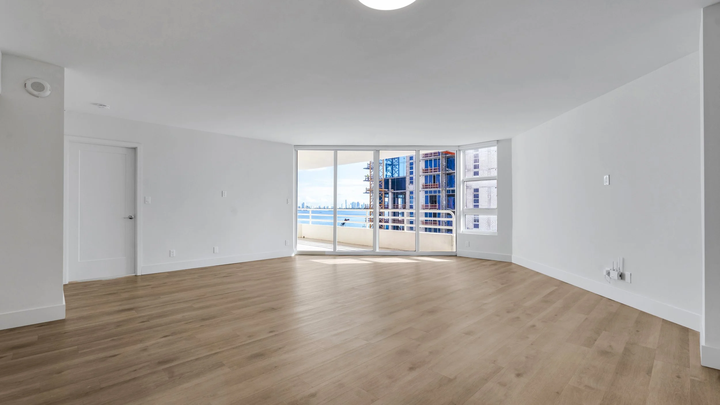Empty living room with white walls, wood flooring, and large glass doors opening to a balcony with city and water view, under a gray ceiling.