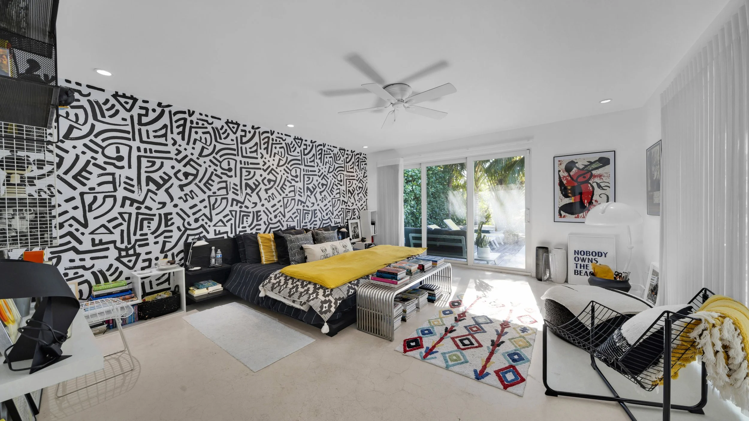 Bright modern living room with geometric black and white accent wall, large glass sliding doors leading to outdoor patio, yellow bedding, various artworks, and contemporary furniture and decor.