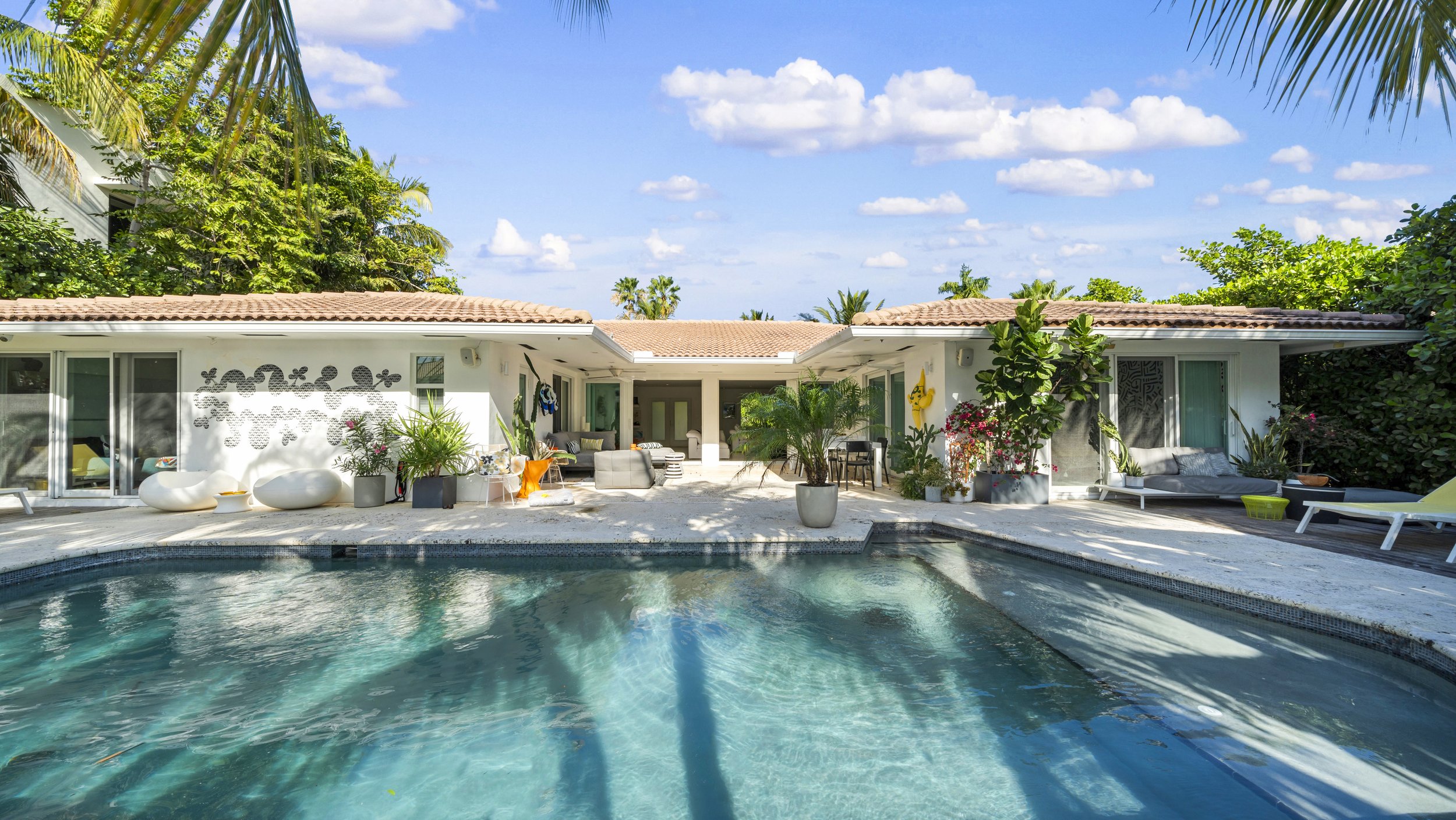A backyard pool area in front of a modern white house with outdoor furniture and lush greenery, under a blue sky with scattered clouds.