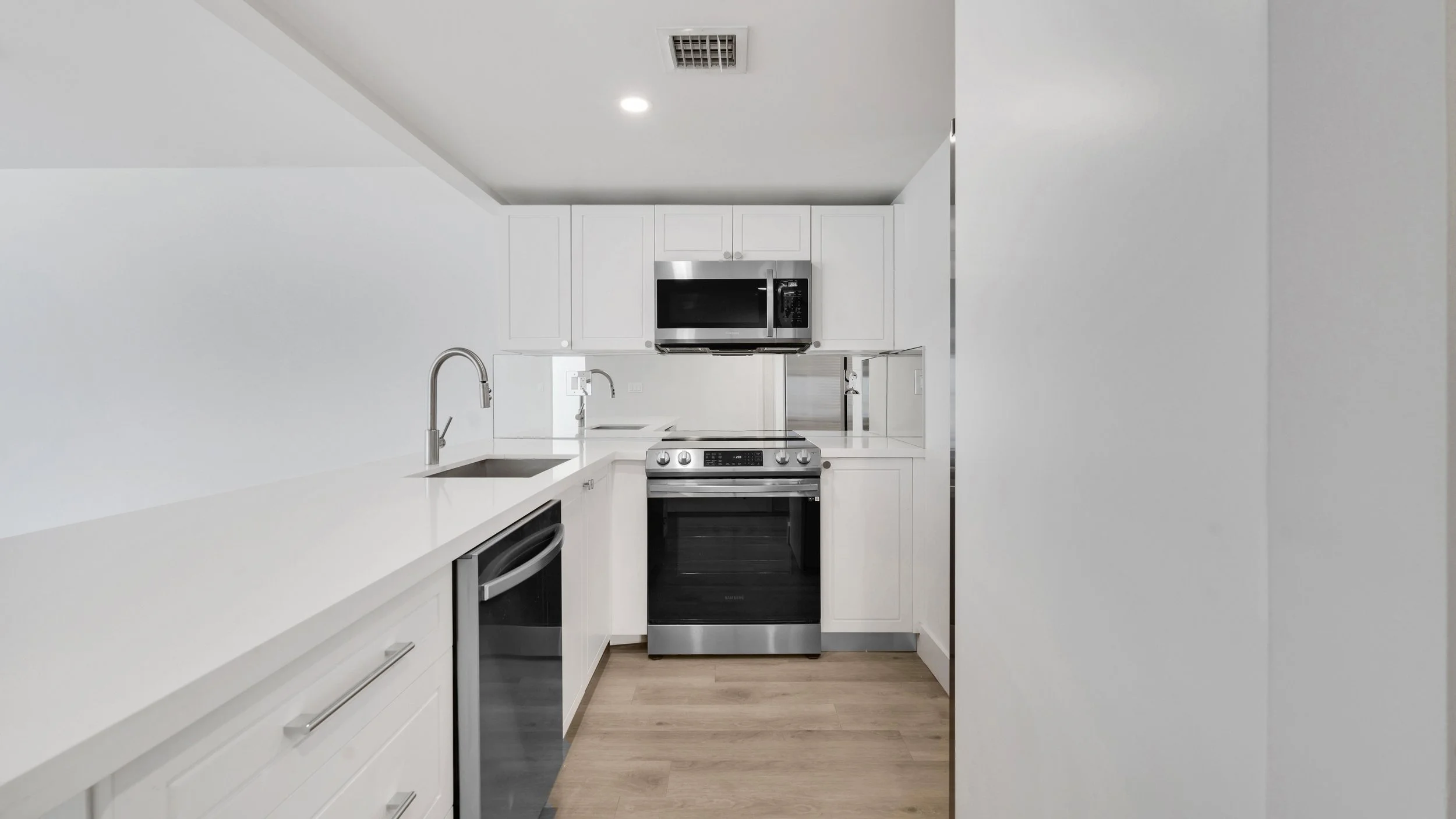 Modern kitchen with white cabinets, stainless steel appliances including a microwave, oven, and dishwasher, with a white countertop and light wooden floor.