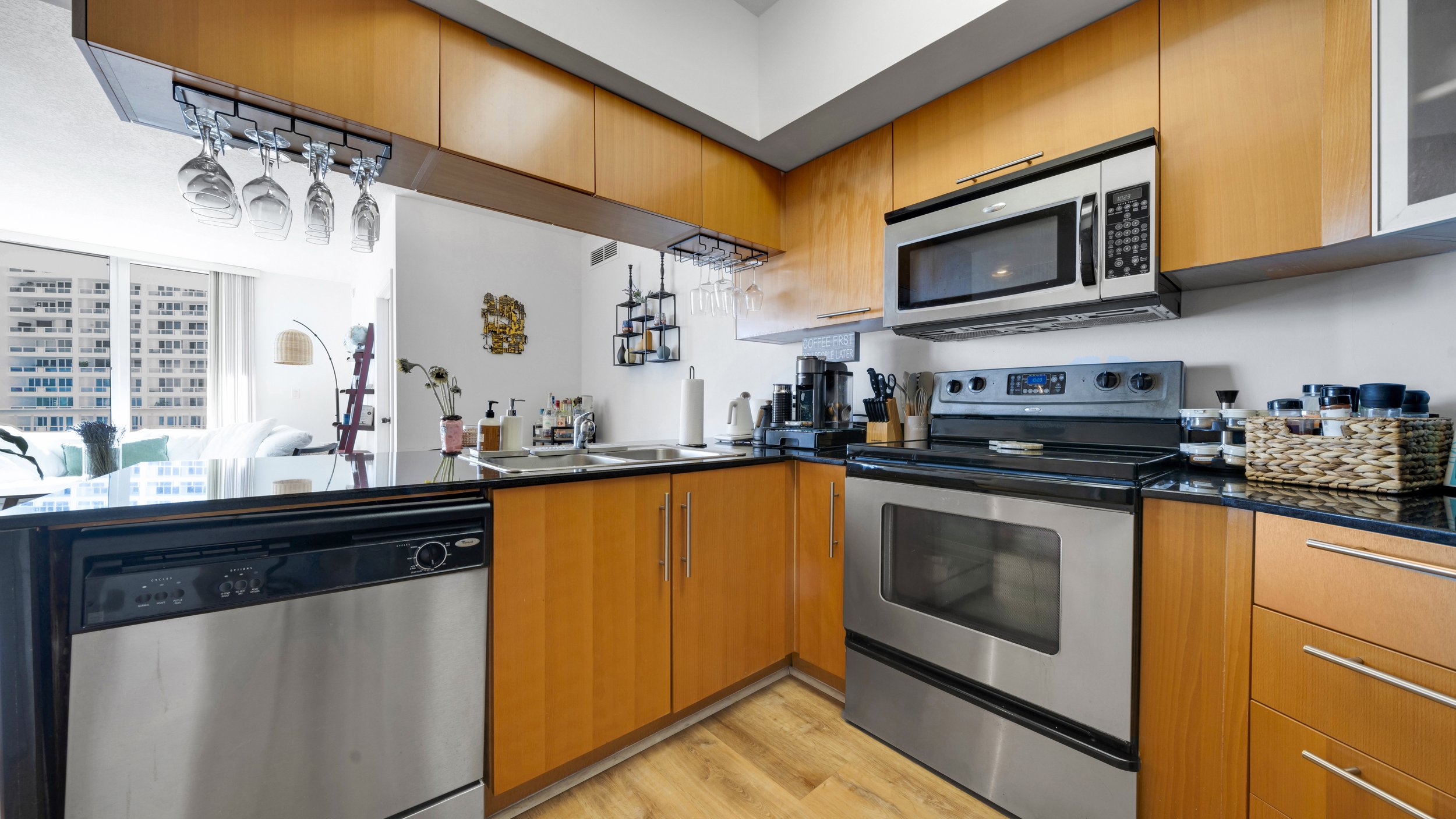 Modern kitchen with wooden cabinets, stainless steel appliances including a microwave, oven, and dishwasher, black countertops, a double sink, and a bar area with hanging glassware, overlooking a bright living space with large windows and city views.