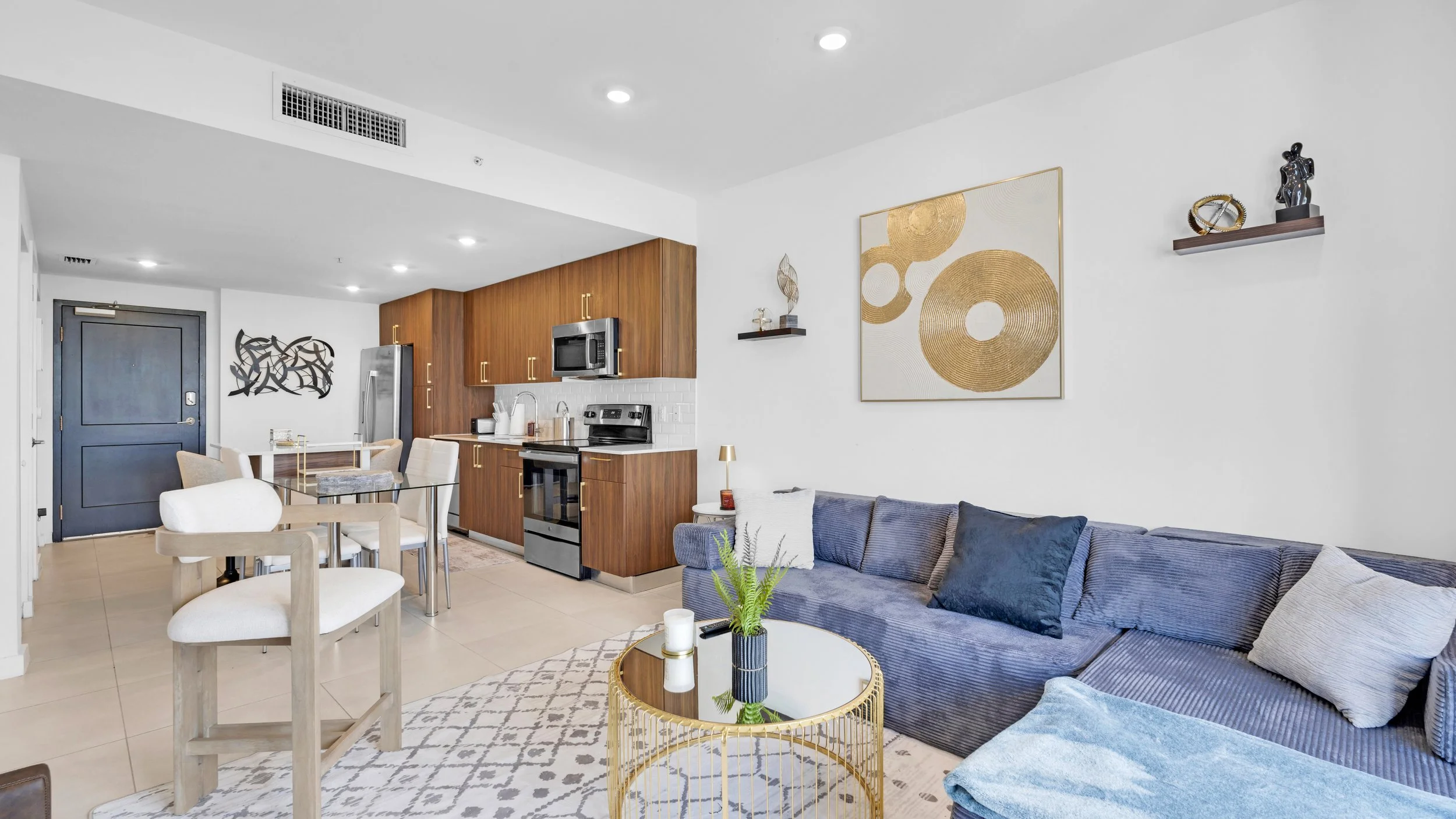 Living room with navy blue sofa, modern glass coffee table with gold frame, beige and white patterned rug, and a corner with wooden kitchen cabinets, stainless steel appliances, and decorative wall art.