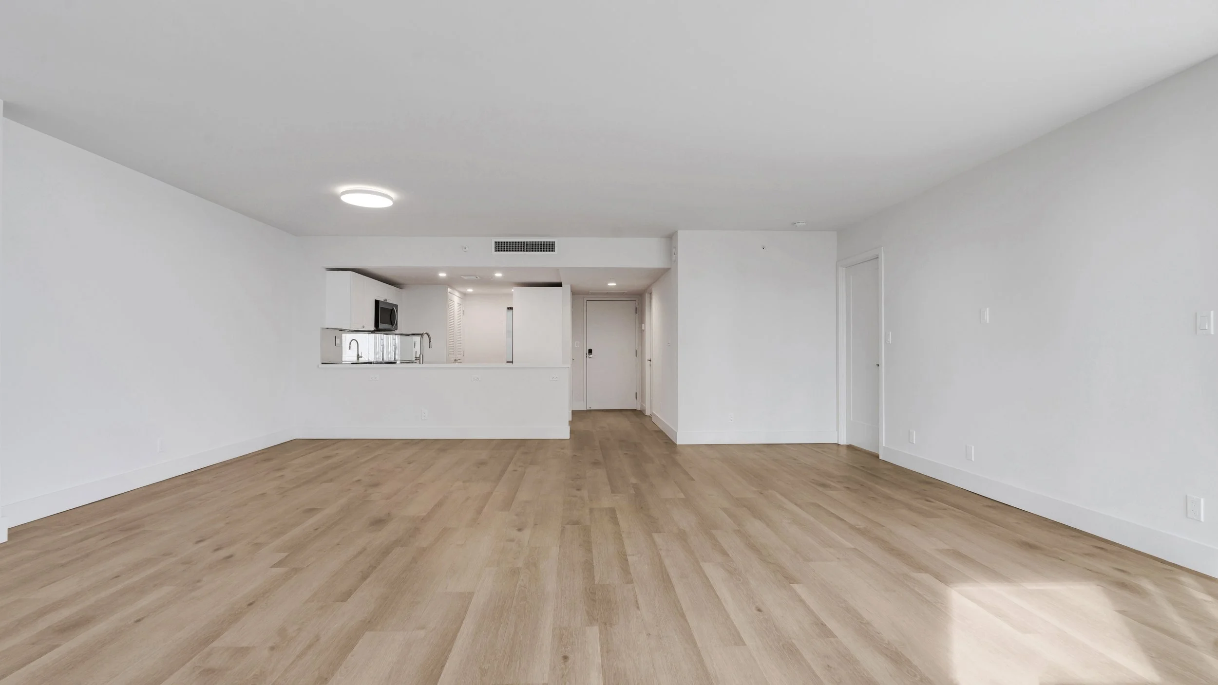 Empty living room with white walls and light wood flooring, open kitchen in the background, hardwood floors and ceiling lights.