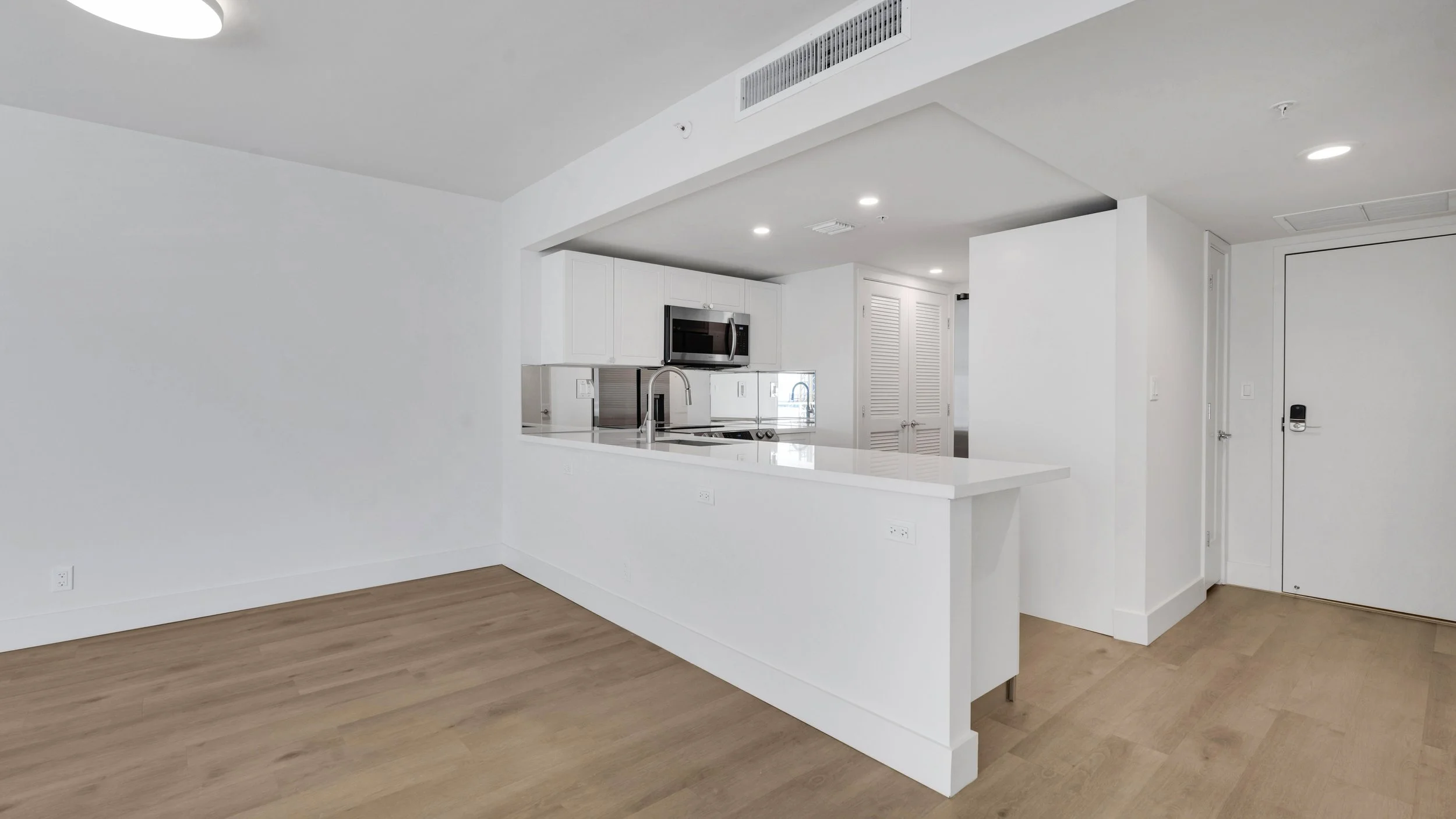 Empty modern kitchen with white cabinets, stainless steel appliances, white countertops, and hardwood floors in an apartment or condo.