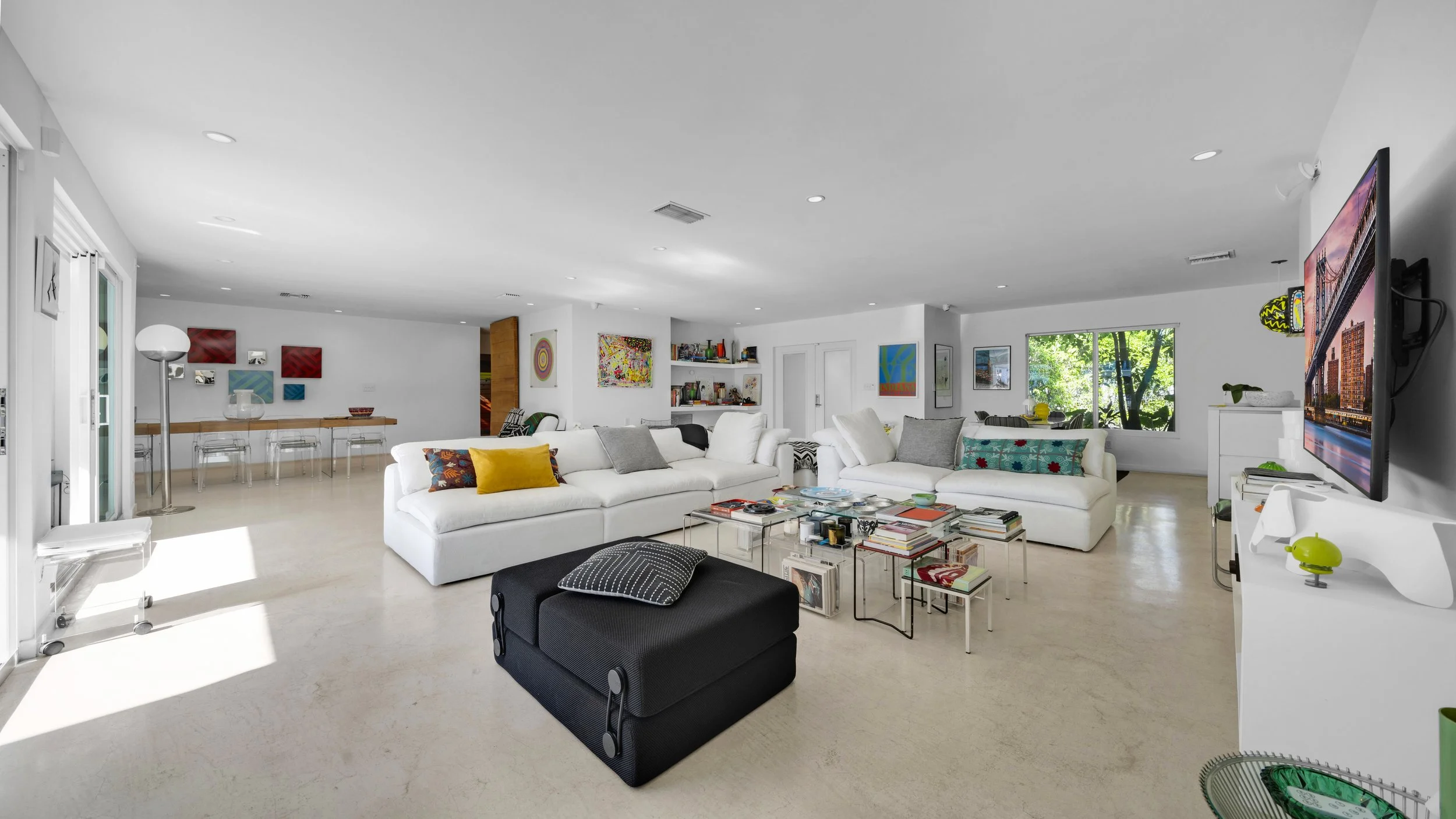 Bright and modern living room with white sofas, colorful pillows, glass coffee tables, wall-mounted TV, and decorated walls with artwork, large windows showing greenery outside, and a dining area in the background.