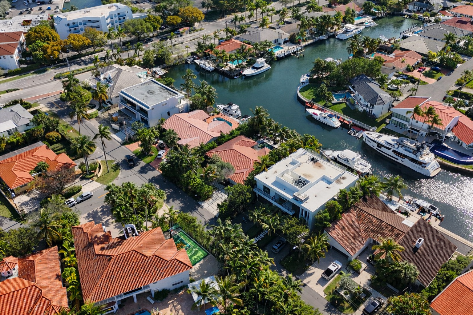 Aerial view of a residential neighborhood with houses, palm trees, and cars lining the streets, along a canal with boats docked.