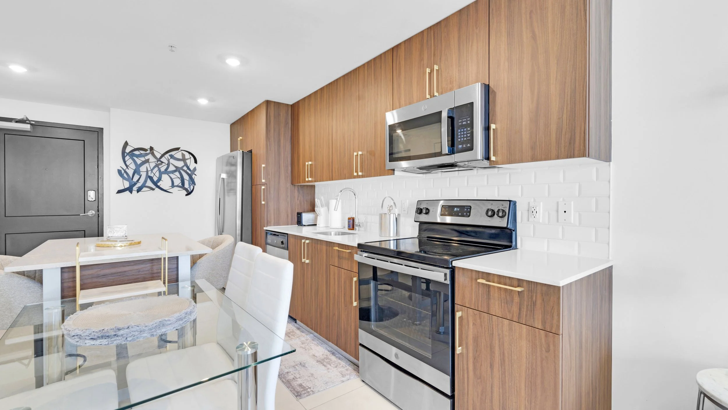 Modern kitchen with wooden cabinets, stainless steel appliances including refrigerator, microwave, and oven, white subway tile backsplash, and a glass dining table with white chairs.