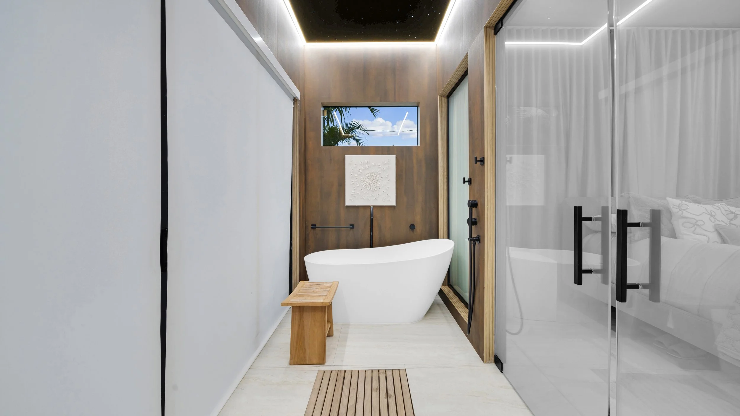 Modern bathroom with a free-standing bathtub, small wooden stool, and a window showing blue sky and palm trees. The room has white walls, a wood accent wall, black fixtures, and sliding glass doors with black handles.