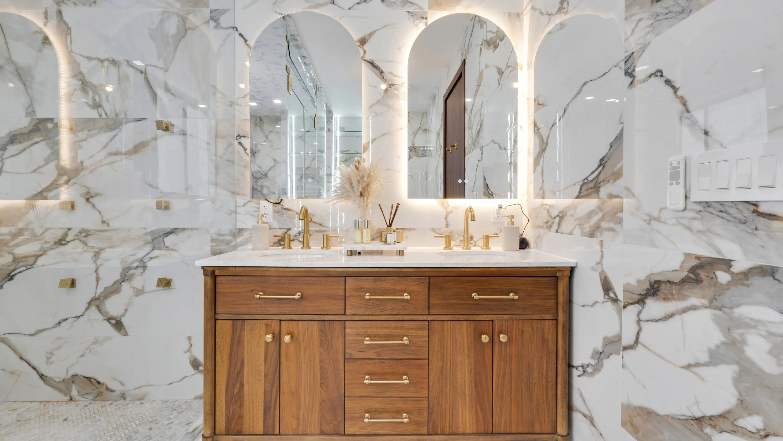 A luxurious bathroom vanity with a marble countertop and backsplash, featuring two gold faucets, a wooden cabinet with gold handles, a decorative tray with reed diffusers, and a mirror with backlit LED lighting on marble walls.