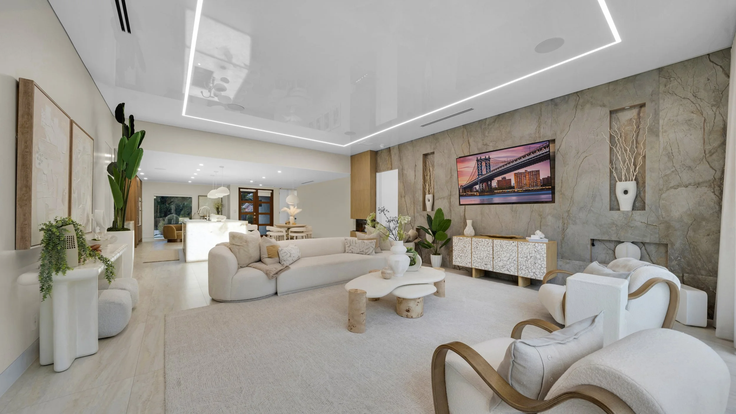 Modern living room with white couches, a large flat-screen TV on a marble accent wall, potted plants, and natural light.
