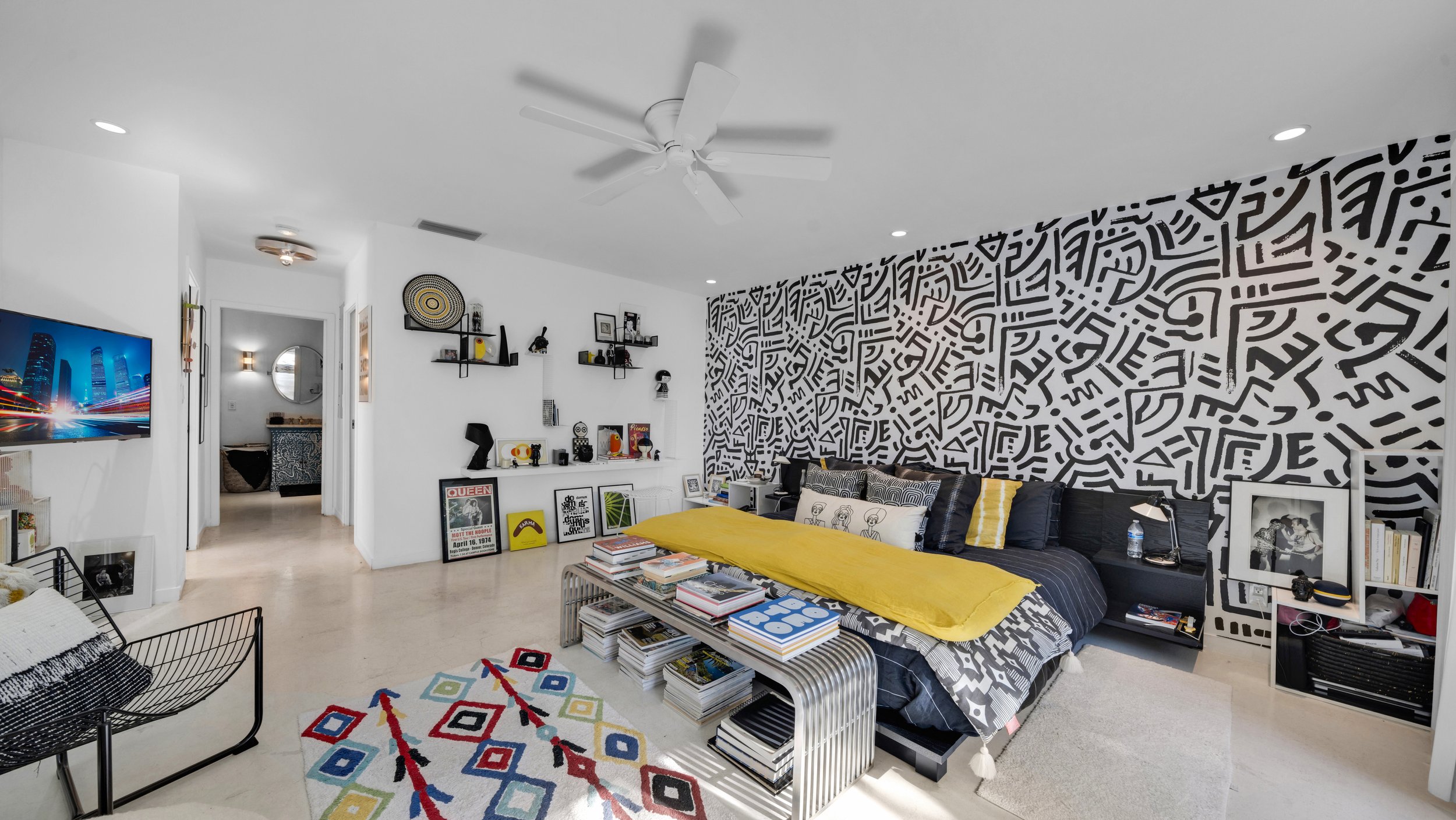 Modern bedroom with black and white abstract patterned wall, yellow blanket on bed, colorful geometric rug, and books and artwork on shelves.