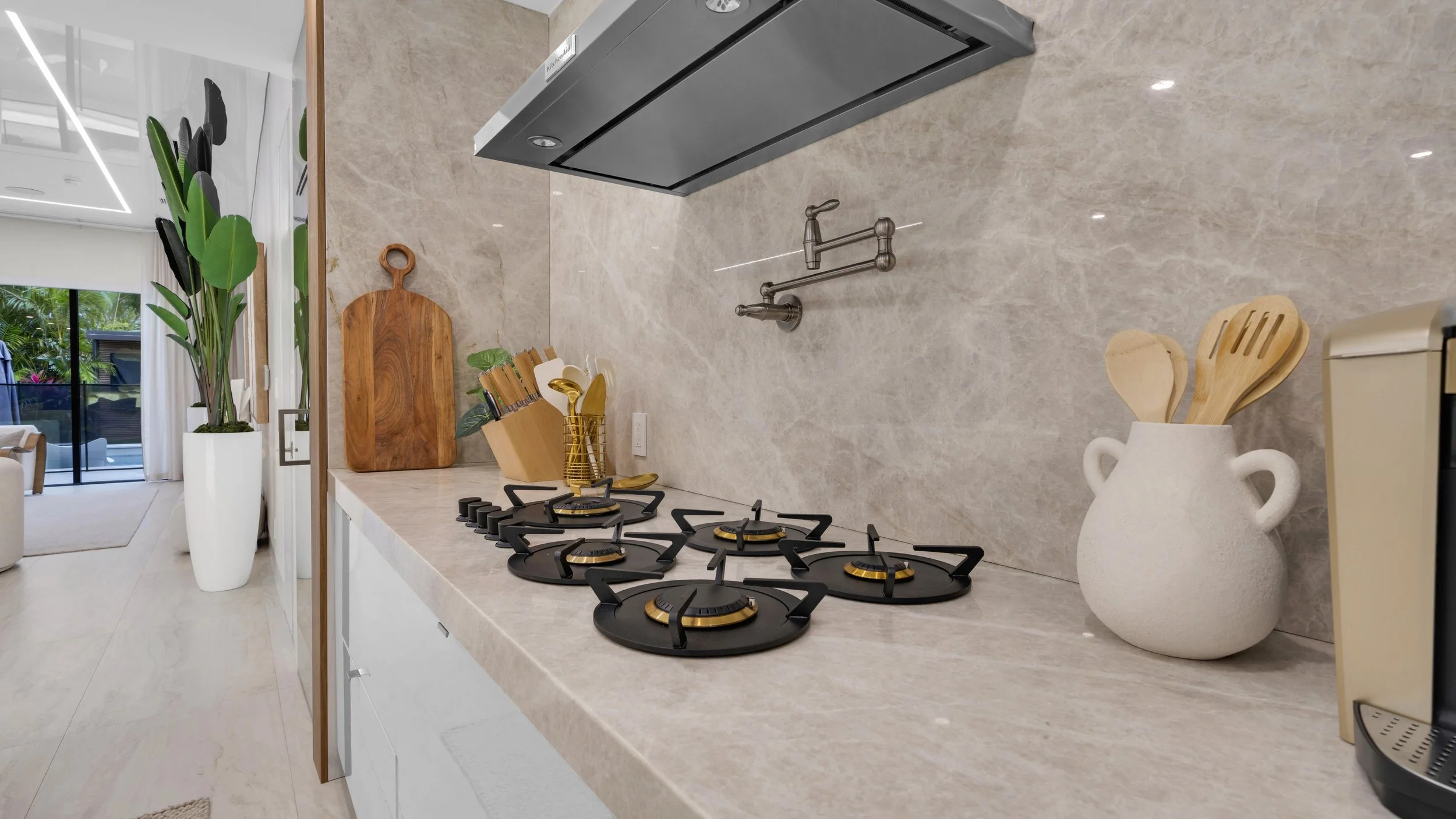 Modern kitchen counter with four black and gold gas burners, a cutting board, a utensil holder with knives, a white vase with wooden spoons, and a container of coffee maker.