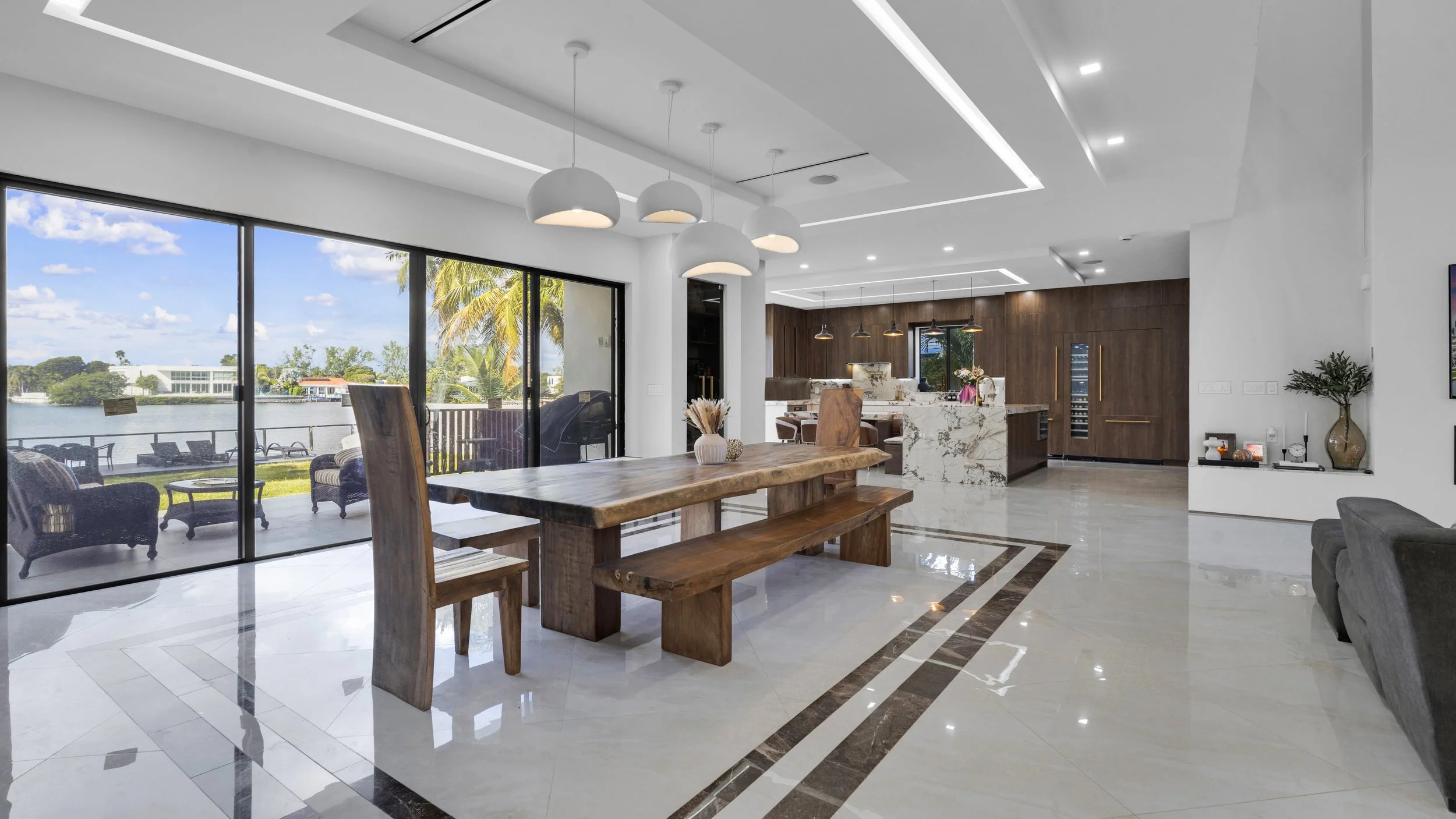 Modern open-concept living space with large glass sliding doors, a wooden dining table, and a kitchen with marble countertops and dark wood cabinets, overlooking a patio and lake.