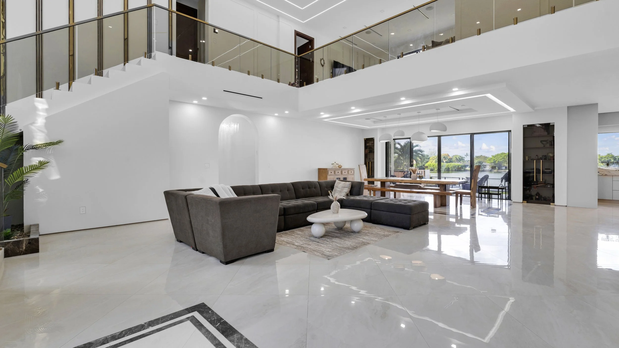 Spacious, modern living room with large sectional sofa, white coffee table, and dining area with wooden table and chairs, with large windows overlooking a lake.