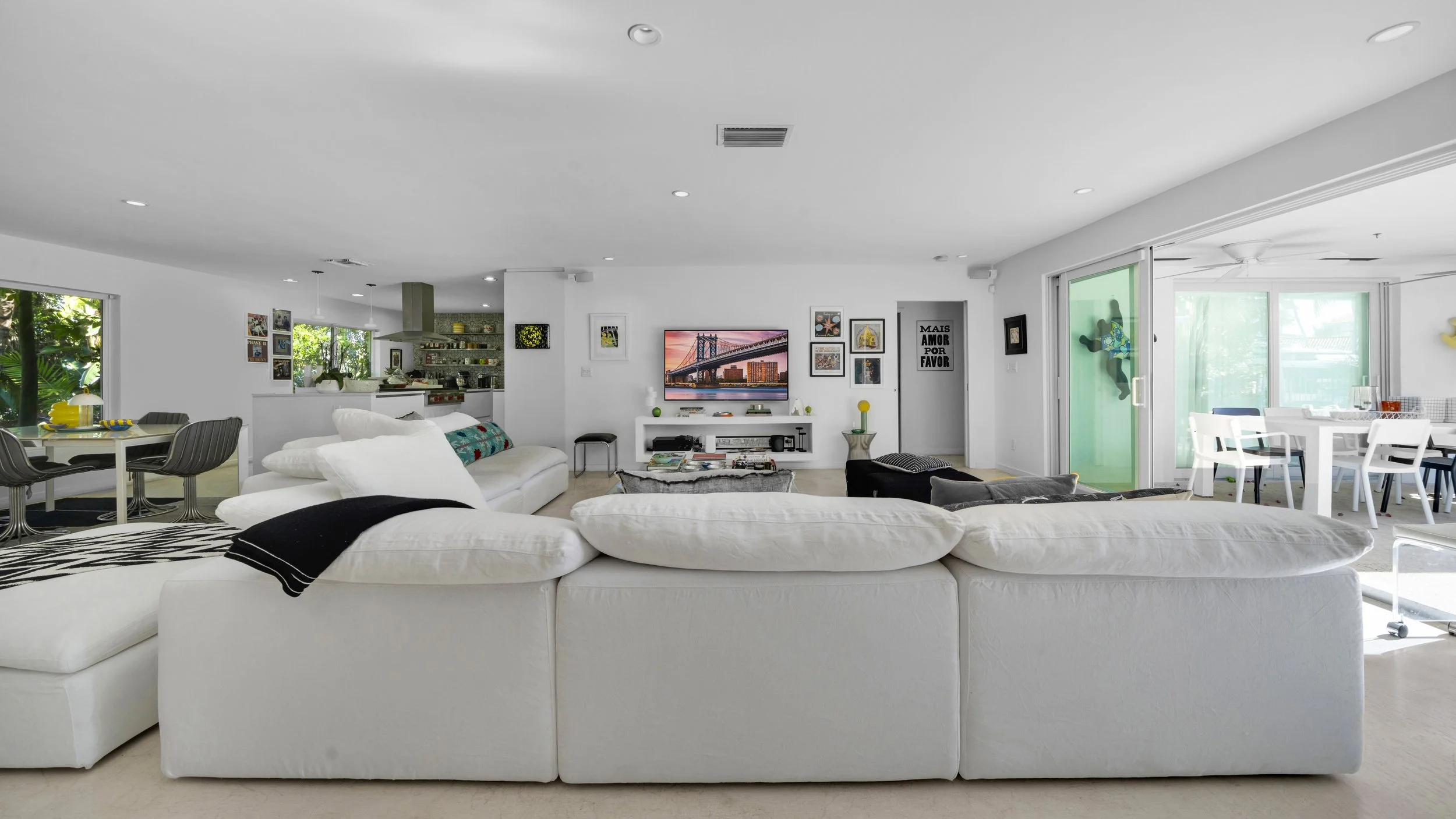 Open-concept living room with white sectional sofa facing a wall-mounted TV, dining area on the right with white table and chairs, and kitchen in the background, all illuminated by natural light from large windows and sliding glass doors.