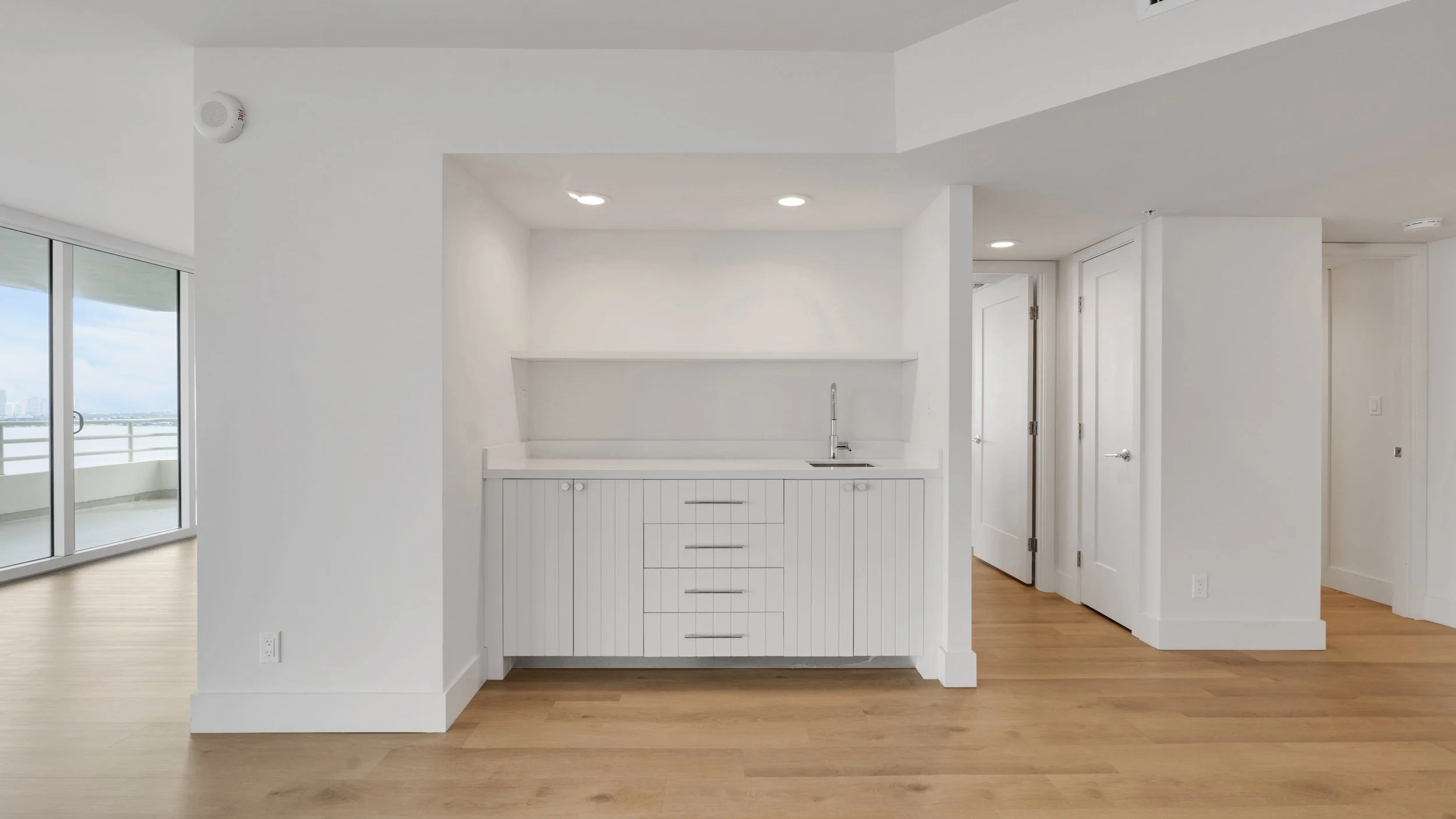 Modern apartment interior with a small white kitchenette, wooden flooring, and a large sliding glass door leading to a balcony with a city view.