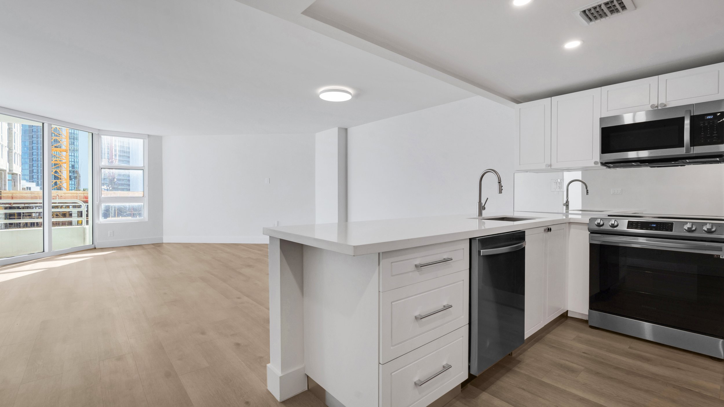 Modern kitchen with white cabinets, stainless steel appliances, and a view of an open living area with large windows showing city buildings.