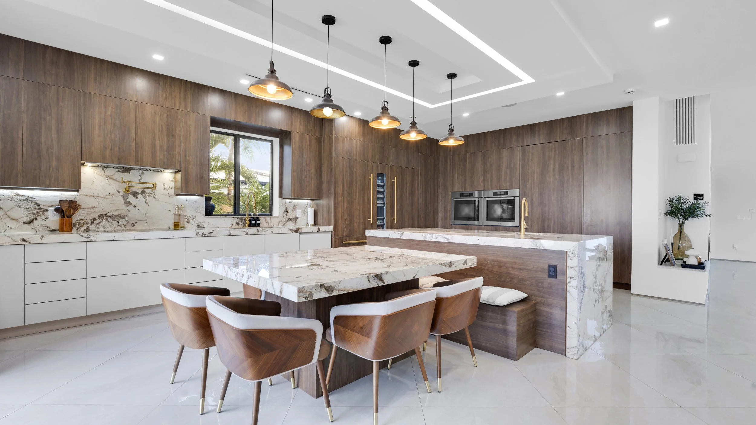 Modern kitchen with white cabinetry, marble countertops, wooden accents, a dining table with matching chairs, black pendant lights, and a window showing outdoor greenery.