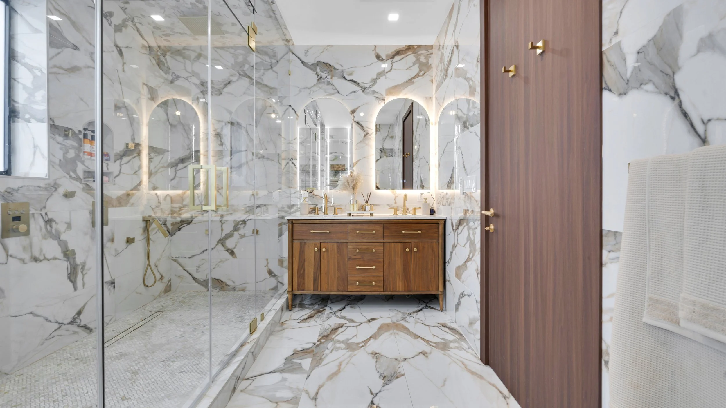 Luxurious bathroom with marble walls and floors, a wooden vanity with gold hardware, illuminated mirrors, and a glass-enclosed shower.