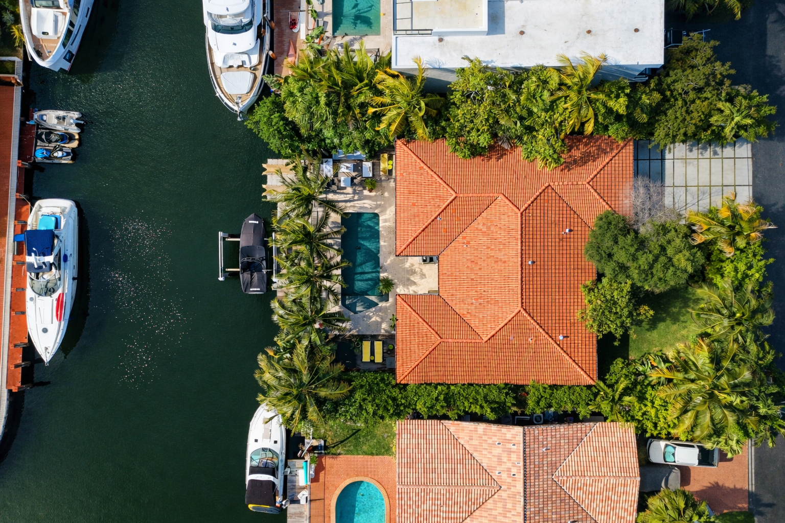Aerial view of a waterfront property with boats docked along a canal, a house with orange tiled roof, a swimming pool, and lush green trees.