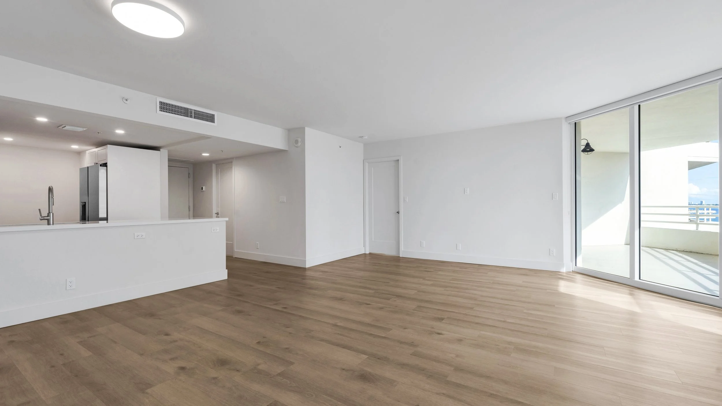 Empty modern apartment living room with wood flooring, white walls, and a large sliding glass door leading to a balcony with city view.