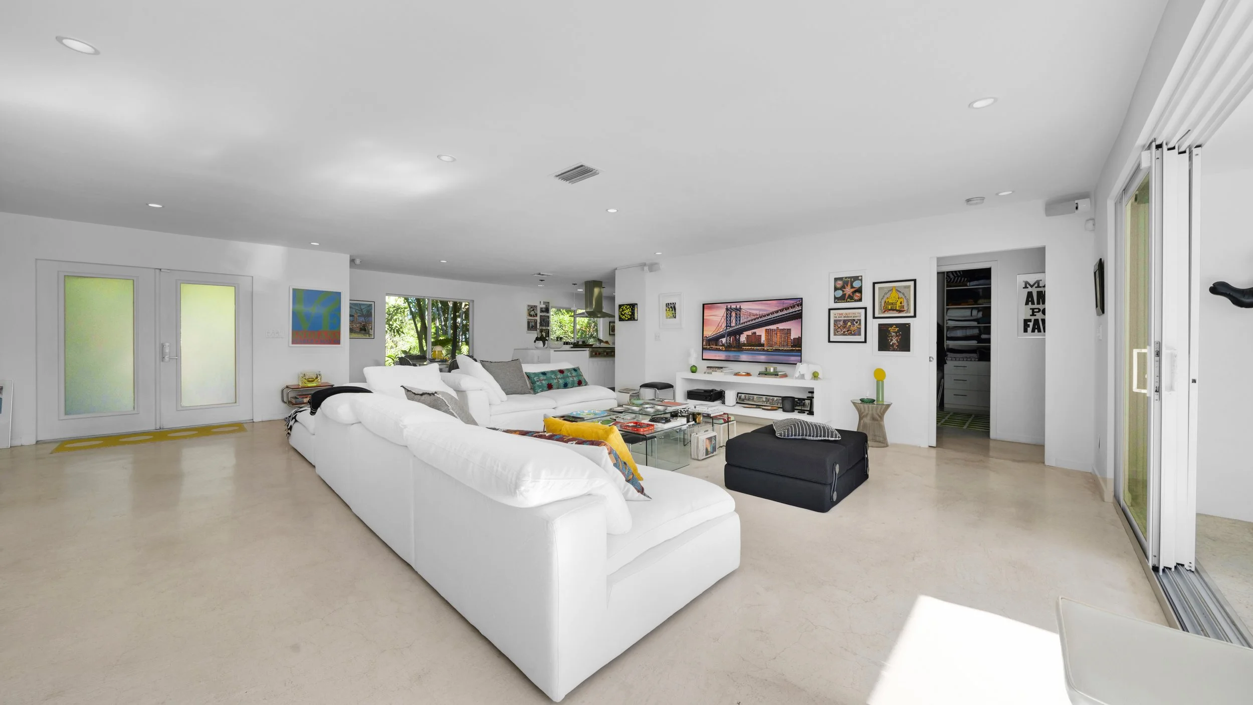 Spacious living room with white couches, a glass coffee table, colorful art on walls, and a TV mounted on a white entertainment center, with large windows and sliding glass doors.