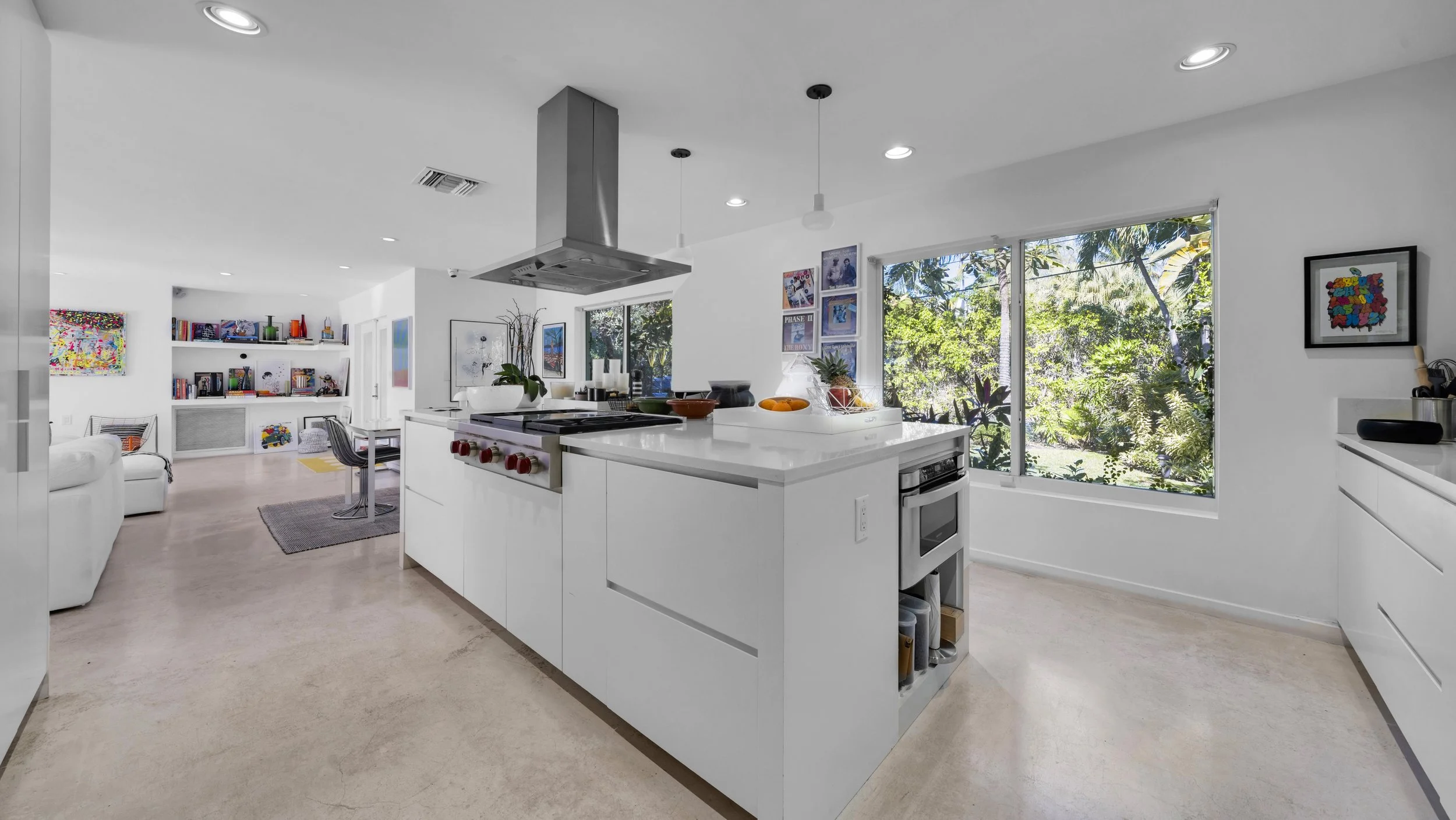 A modern kitchen with white cabinets, a central island, large windows with a view of greenery, and a white living area with artwork and books.