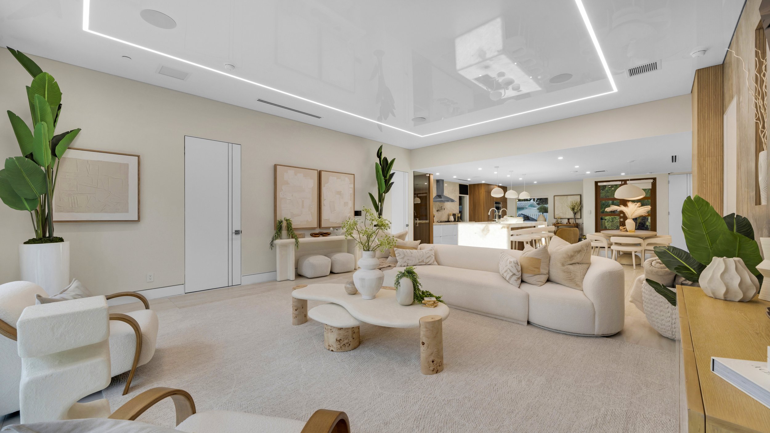 Modern open-concept living room with white sectional sofa, light-colored armchair, wood accents, potted plants, and minimalistic decor, seamlessly connected to a kitchen and dining area.
