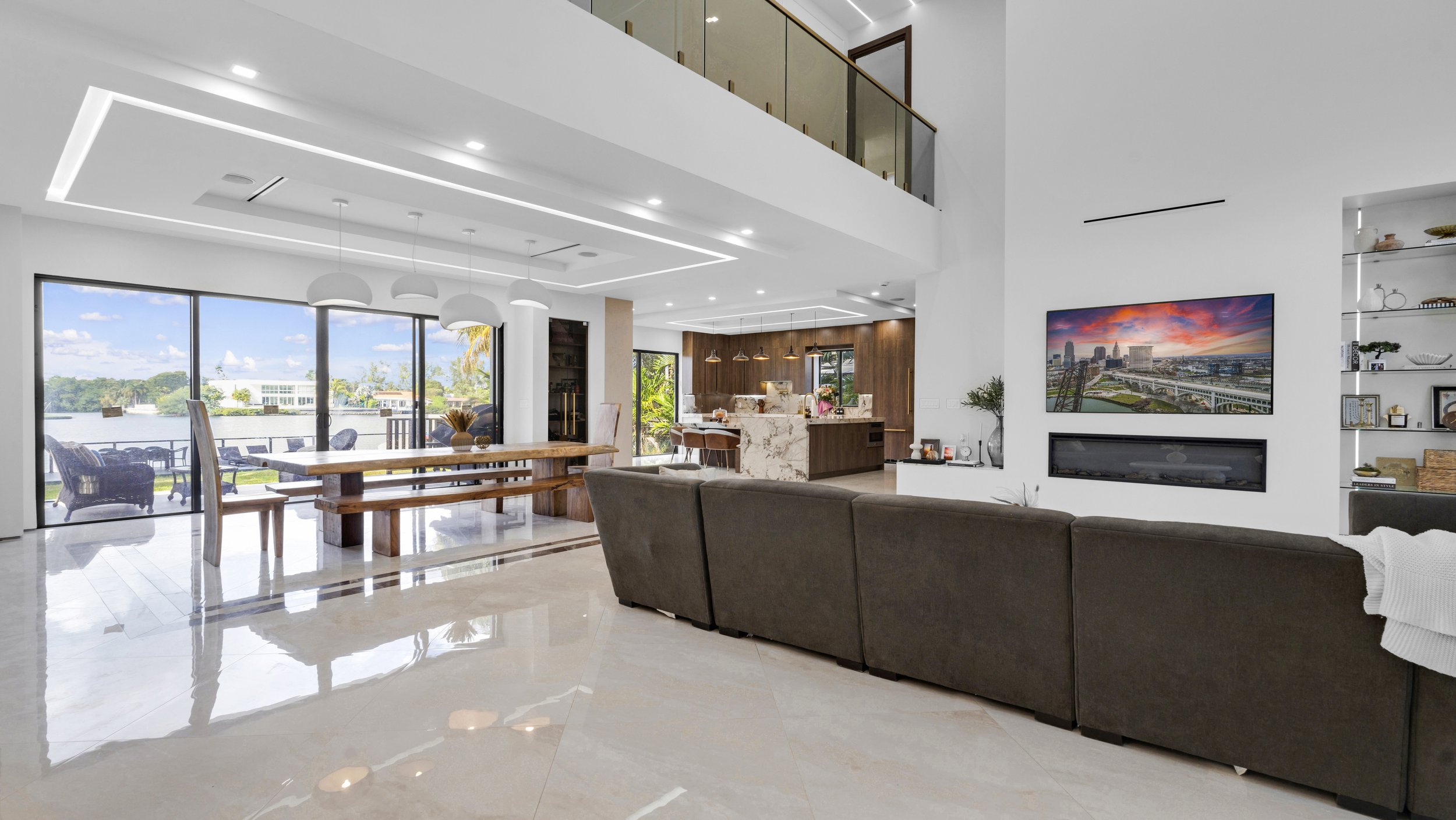 Modern living room with large glass sliding doors opening to an outdoor seating area and water view, a brown sectional sofa facing an electric fireplace and wall-mounted TV, open kitchen with marble island, and contemporary decor.
